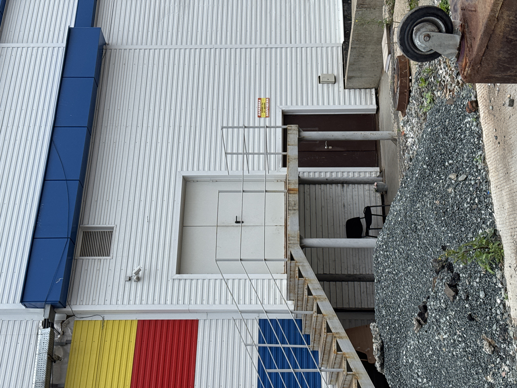 This image depicts the exterior of a building, likely an industrial or commercial facility, in Izhevsk, Russia. The building features a white corrugated metal facade. A section on the left side of the building is vertically divided into three colored bands: yellow at the top, red in the middle, and blue at the bottom. Above the white facade, there is a blue horizontal structure. A security camera is mounted on the white wall.
There are two entrances visible. The primary entrance on the right is a double white door accessed by a metal staircase with a handrail. To the right of this staircase, there is another brown door. A yellow sign with red lettering, which translates to "PARKING PROHIBITED FROM THE BUILDING," is affixed to the white wall between the two doors.
In front of the building, a large pile of grey gravel and construction debris is present. A black office chair is positioned under the staircase. The ground is a mix of concrete and gravel. In the foreground, a rusty metal object with a wheel attached is visible, along with some scattered debris and small plants. The scene appears to be outdoors, under an overcast sky.