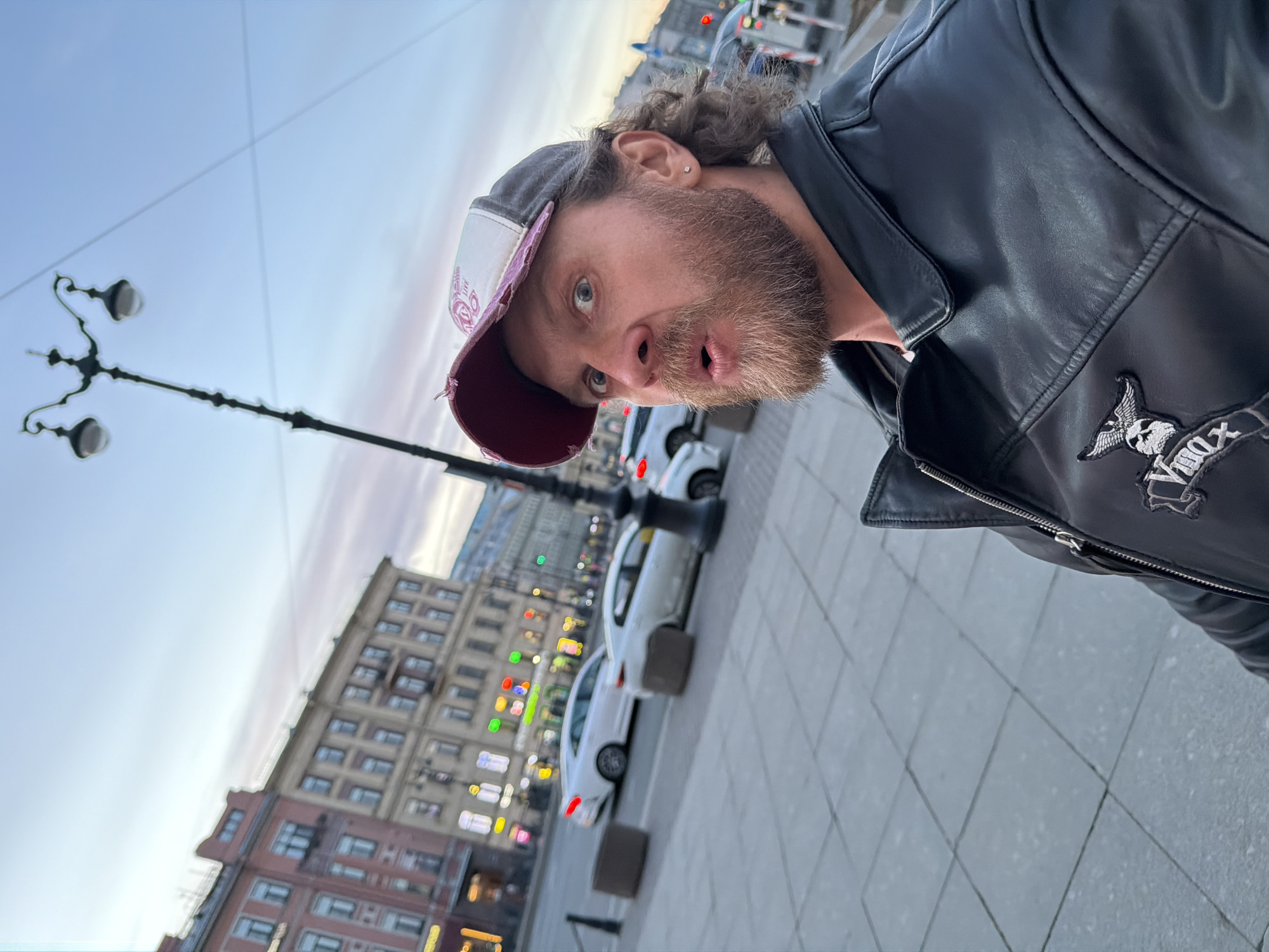 A man is seen from a skewed, low-angle perspective, potentially a selfie. He is wearing a black leather jacket with a patch of a skull-and-wings motif, and a worn baseball cap with a distressed brim. He has a beard, wavy hair, and a surprised expression.
The background is an urban streetscape, featuring multi-story buildings with illuminated windows, streetlights, and parked cars. The presence of the architectural style suggests this could be Saint Petersburg, Russia. The buildings are brick and stone. The sky is a gradient of pale colors.