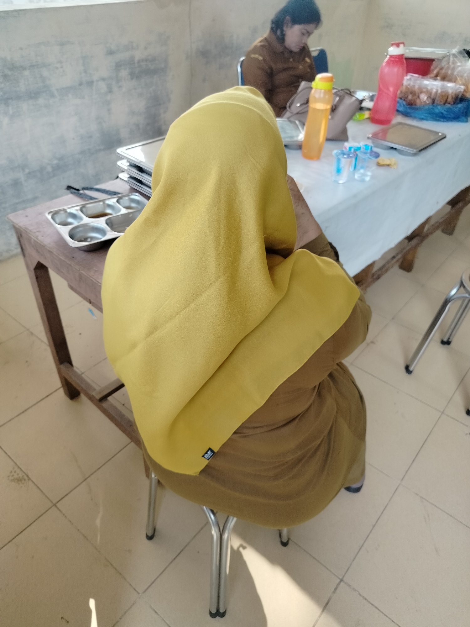 In an indoor setting that appears to be a cafeteria or break room, two women are seated at a table. The woman in the foreground, viewed from behind, wears a yellow hijab and a brown uniform, and is sitting on a metal stool. In front of her on a wooden table is a compartmentalized metal tray with food. To her right, a table covered with a white cloth is laden with items including two water bottles, one orange and one red, and clear plastic cups. Another woman, wearing a similar brown uniform, is visible in the background, seated at the same table and looking down. The floor is tiled with light-colored square tiles. The wall behind the women is textured and light-colored, with some darker markings. This scene likely takes place in Purwodadi, Indonesia, possibly within a government or educational institution given the uniforms.