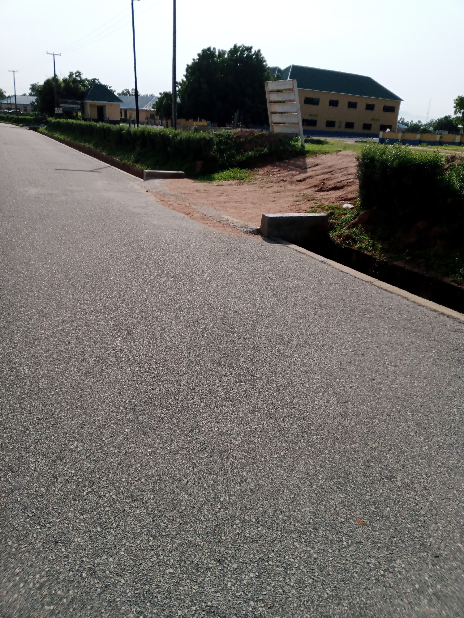 The image shows a paved road in Jalingo, Nigeria. On the left side of the road, there is a building with a green roof and yellow walls. Behind the building, there are trees. On the right side of the road, there is a larger yellow building with a green roof and blue trim. In front of this building, there is a sandy area with some bushes. There is also a concrete drainage ditch along the side of the road. The sky is overcast.