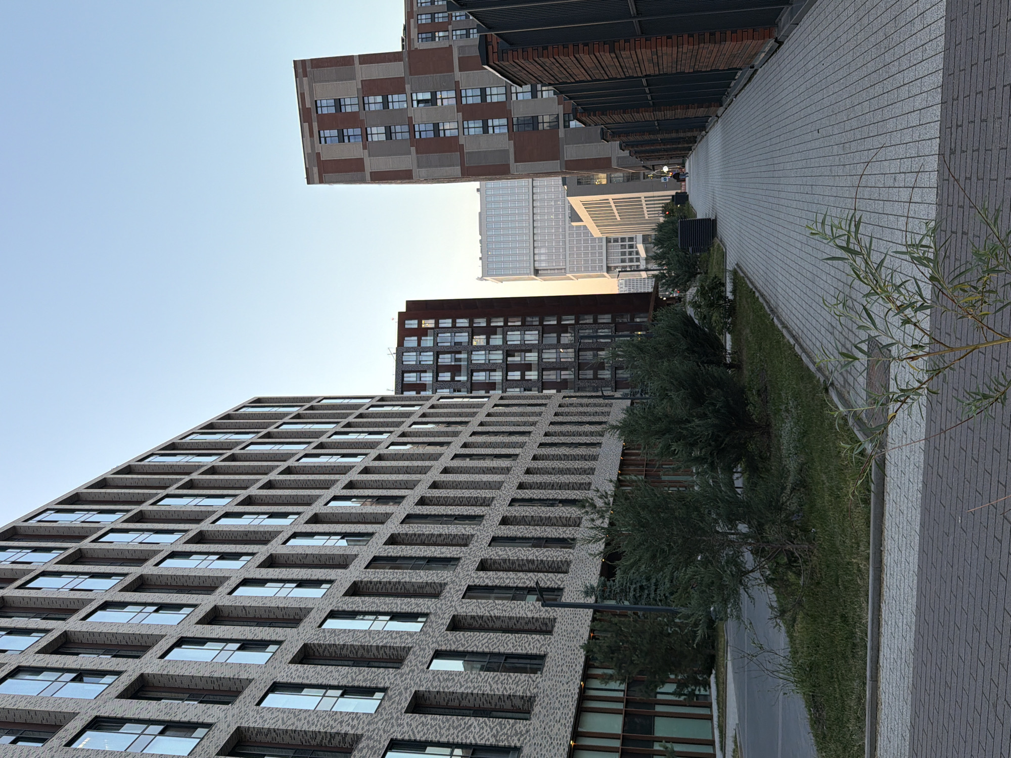The image depicts a street view in Moscow, Russia, featuring modern multi-story buildings and a pedestrian walkway. A large, light-colored brick building with numerous windows dominates the left side of the frame. In the background, several other contemporary buildings of varying heights and architectural styles are visible, including a tall brick-colored tower and a lighter, more translucent structure. The walkway is paved with light-colored tiles and is lined with green bushes and trees. A black fence borders the right side of the walkway, constructed from brick and metal. A single person is walking in the distance on the walkway. The sky is clear and pale blue, indicating it might be early morning or late afternoon.