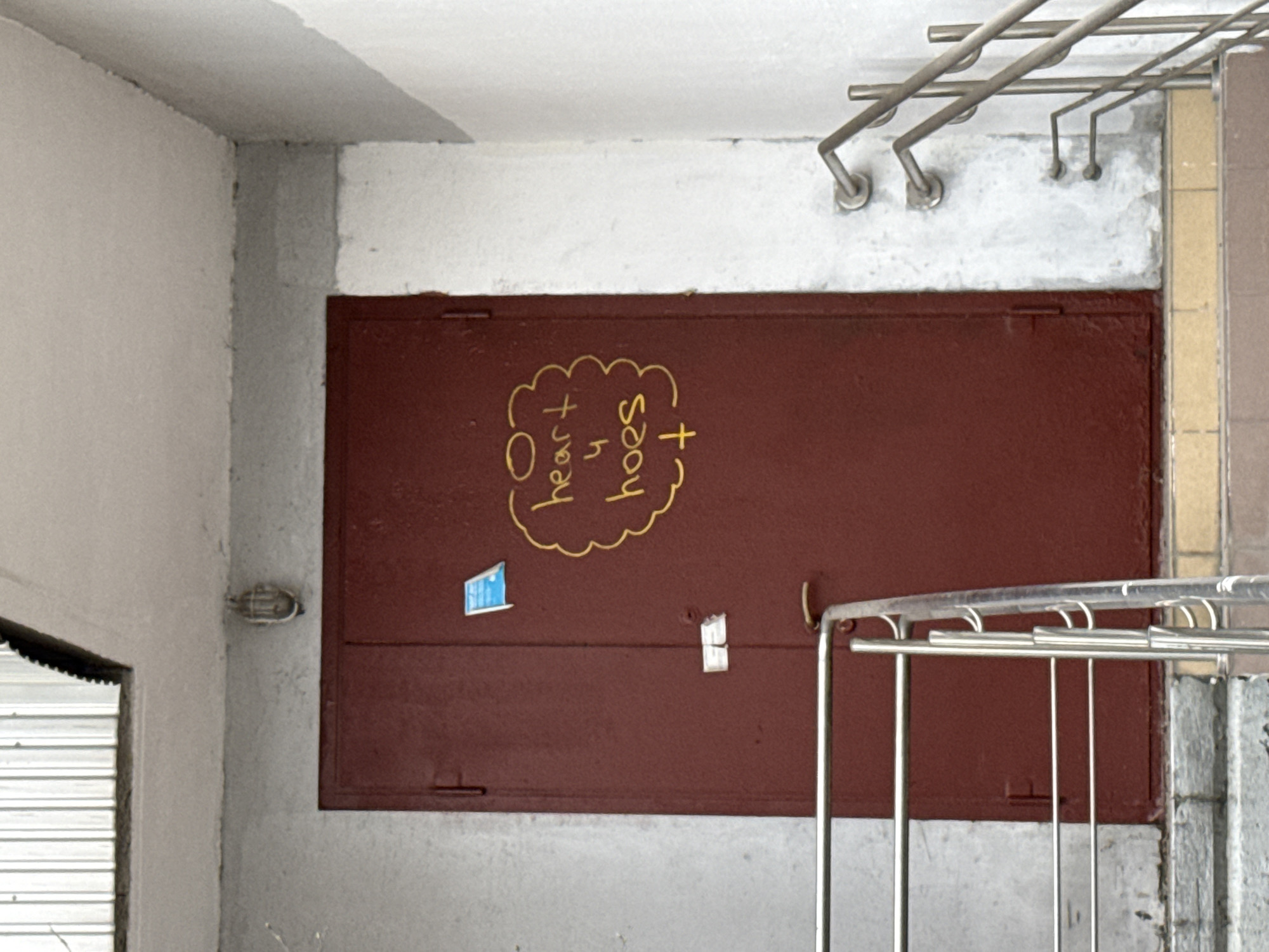The image depicts a stairwell in Moscow, Russia. The walls are painted in light gray and white. A large, maroon-colored metal door occupies a central position in the frame, with graffiti written in yellow paint on it, showing "heart n soul". A small blue sticker is on the left side of the door. A metal handrail is visible on the right side of the door, indicating the presence of stairs. Above the door is a metal clothes drying rack attached to the ceiling. There is a garage door on the left side of the scene.