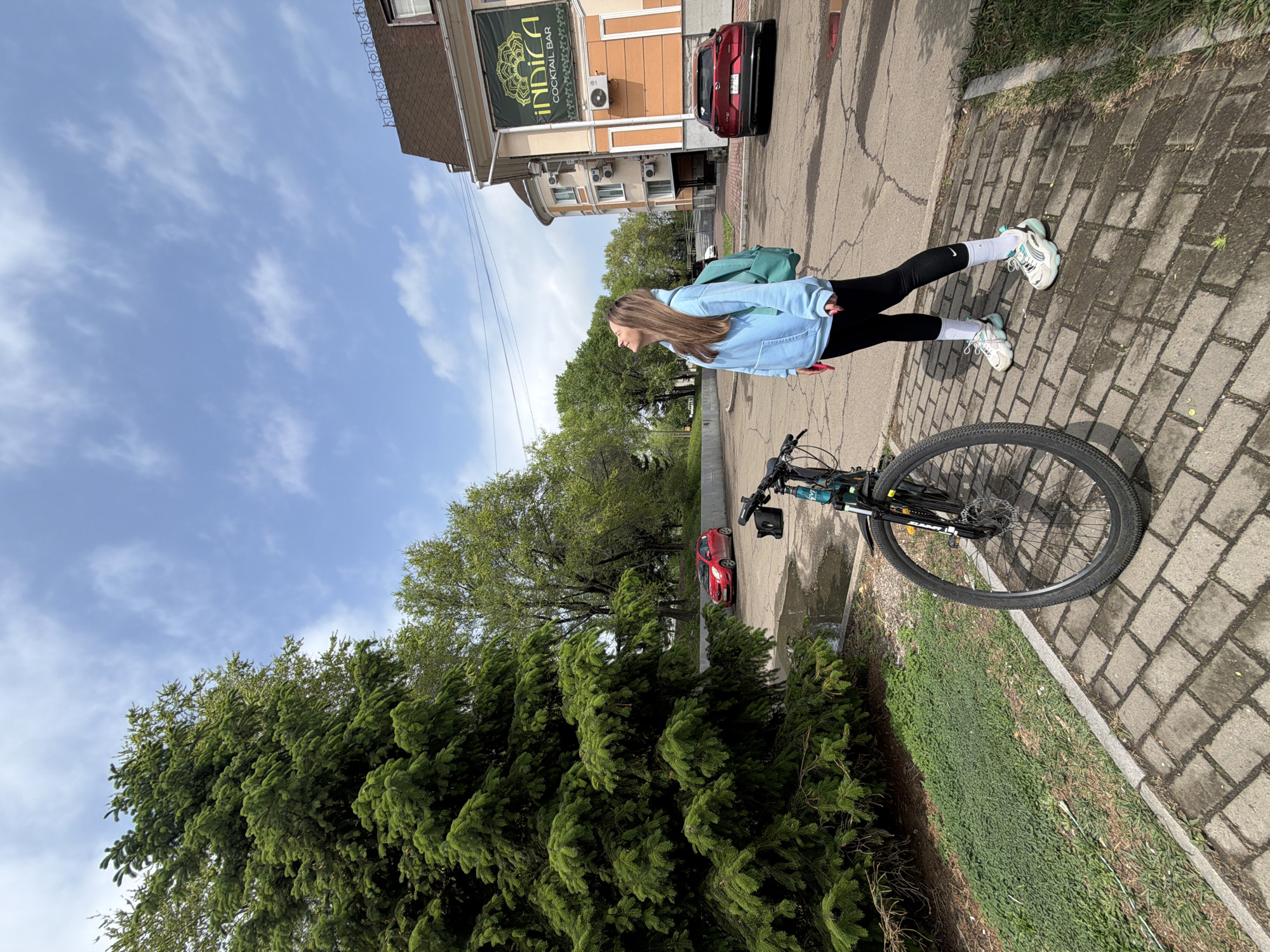 A young woman with long blonde hair stands on a brick sidewalk, a bicycle beside her. She wears a light blue sweatshirt, black leggings, white socks, and white sneakers. A green backpack is slung over her shoulder. Behind her is a building with a sign that reads "INDICA COCKTAIL BAR". A red car is parked in front of the building. In the background are trees, more buildings, and a blue sky with clouds. A coniferous tree is in the lower left corner. Another red car is parked nearby on the street. The location is likely in Birobidzhan, Russia, based on the text in the image.