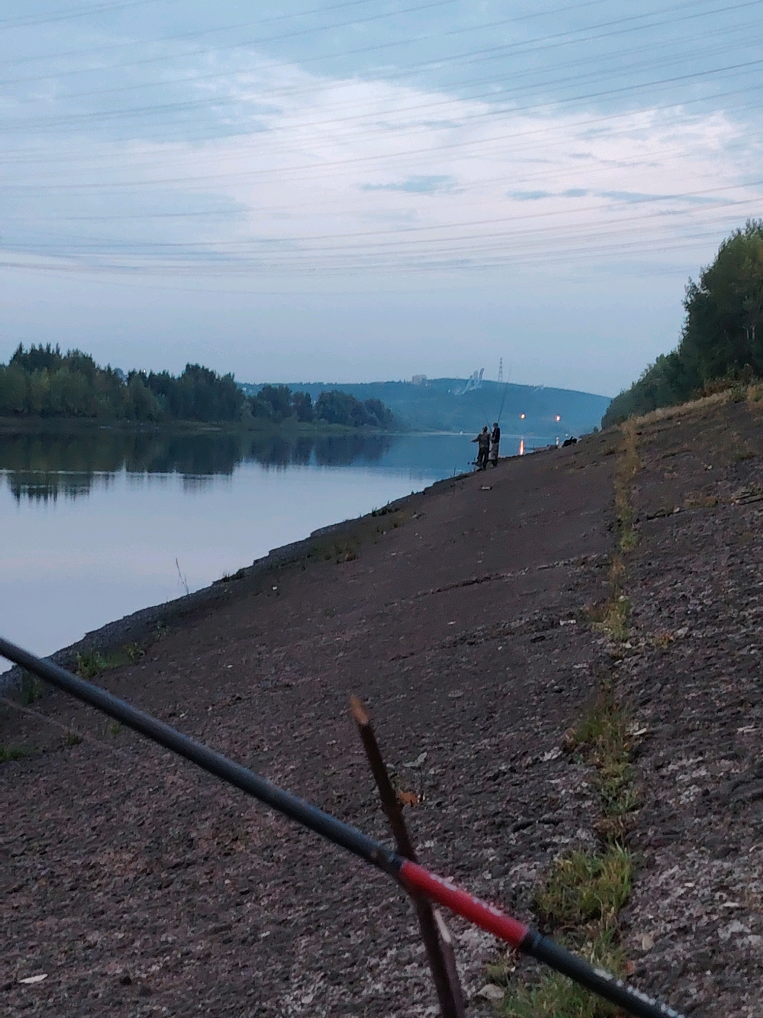 The image depicts a scene on the bank of the Kama River in Chaikovsky, Russia, during what appears to be twilight. In the foreground, a fishing rod with a red and black handle lies angled across the frame, resting on a gravelly, sloped embankment. Further up the embankment, two individuals are visible fishing, casting their lines into the calm river. The river's surface reflects the overcast sky and the distant treeline on the opposite bank. In the background, industrial structures and power lines are faintly visible on a hill. The overall atmosphere is serene, with the soft, diffused light of dusk.