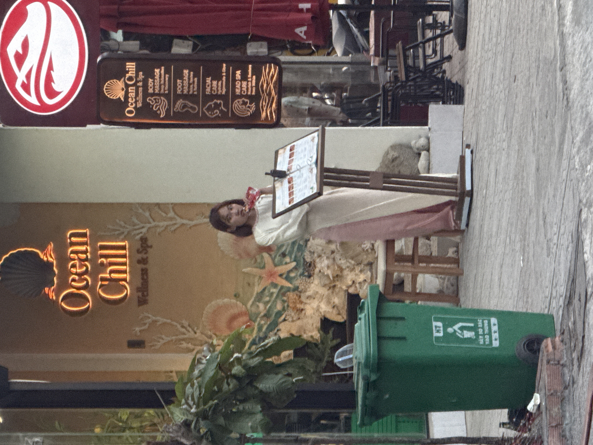 A young woman in a white and pink dress stands next to a menu stand outside a business called "Ocean Chill Wellness & Spa" in Bac Nha Trang Ward, Vietnam. The woman is holding a snack and appears to be looking at the menu. The facade of the spa features decorative shells and coral. To the left of the woman is a green trash can with Vietnamese text and a recycling symbol. In the background, there are outdoor tables and chairs, as well as a red umbrella. The overall scene suggests a street-level commercial area.