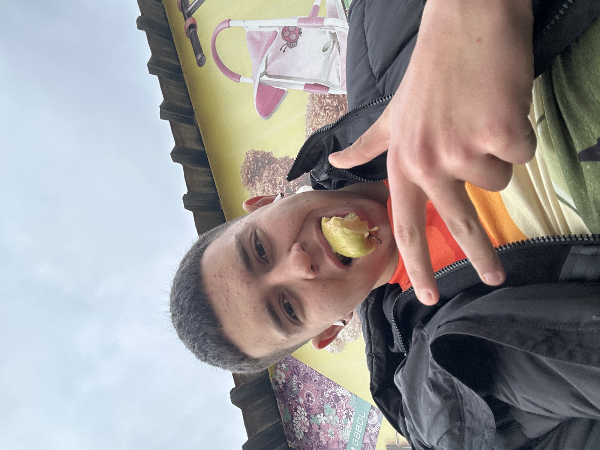 A selfie taken in Zlatograd, Bulgaria, features a young man with short dark hair eating a light-colored fruit, possibly a pear, while making a hand gesture. He wears a black jacket over an orange and tan colored shirt. Behind him is a yellow exterior wall with a painted mural depicting a pink toy stroller and other motifs. The sky is overcast. A patterned item with the word "GABOL" is visible. The building has a dark-colored roof with a saw-tooth edge.