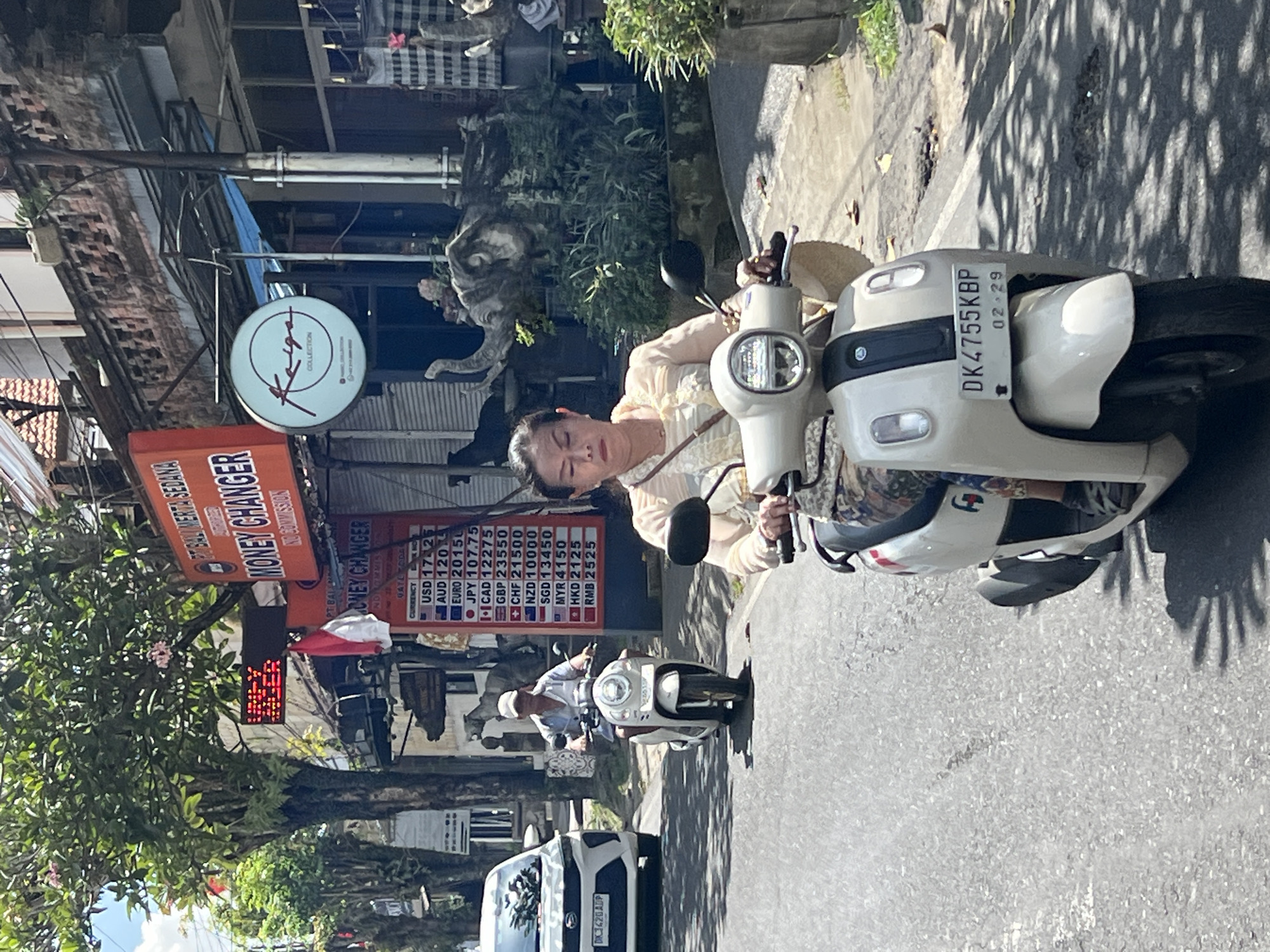 A woman rides a white scooter on a paved road in Ubud District, Indonesia. She is wearing a light-colored blouse and patterned pants. The scooter has a license plate that reads "DK4755KBP 02-29". In the background, a man on another scooter is visible, as is a white car. Signs for a money changer are also present, displaying currency exchange rates. Elephant statues and lush greenery are seen in the background. The scene appears to be a street in a bustling area with shops and trees lining the road.