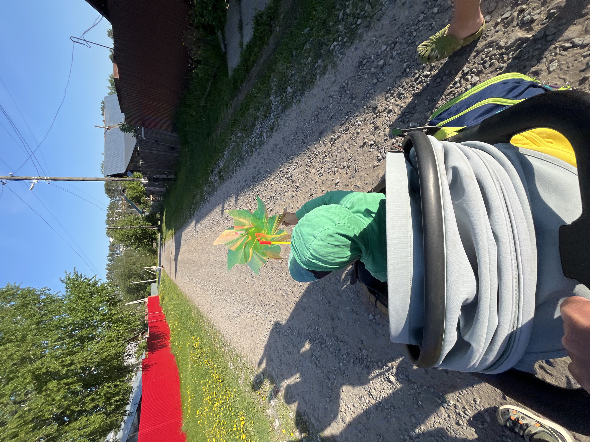 A person, viewed from the perspective of someone pushing a stroller, is on a dirt road in Chaikovsky, Russia, based on the context clues. The person, dressed in a green shirt and cap, is holding a colorful pinwheel. The dirt road is lined with grass and dandelions. A red fence is visible on the left side of the road, along with a cluster of trees. A building with a brown roof is also present on the left side of the road, further down the path. The stroller is visible in the foreground. Another person's foot, clad in a green sandal, is also visible near the top of the image.