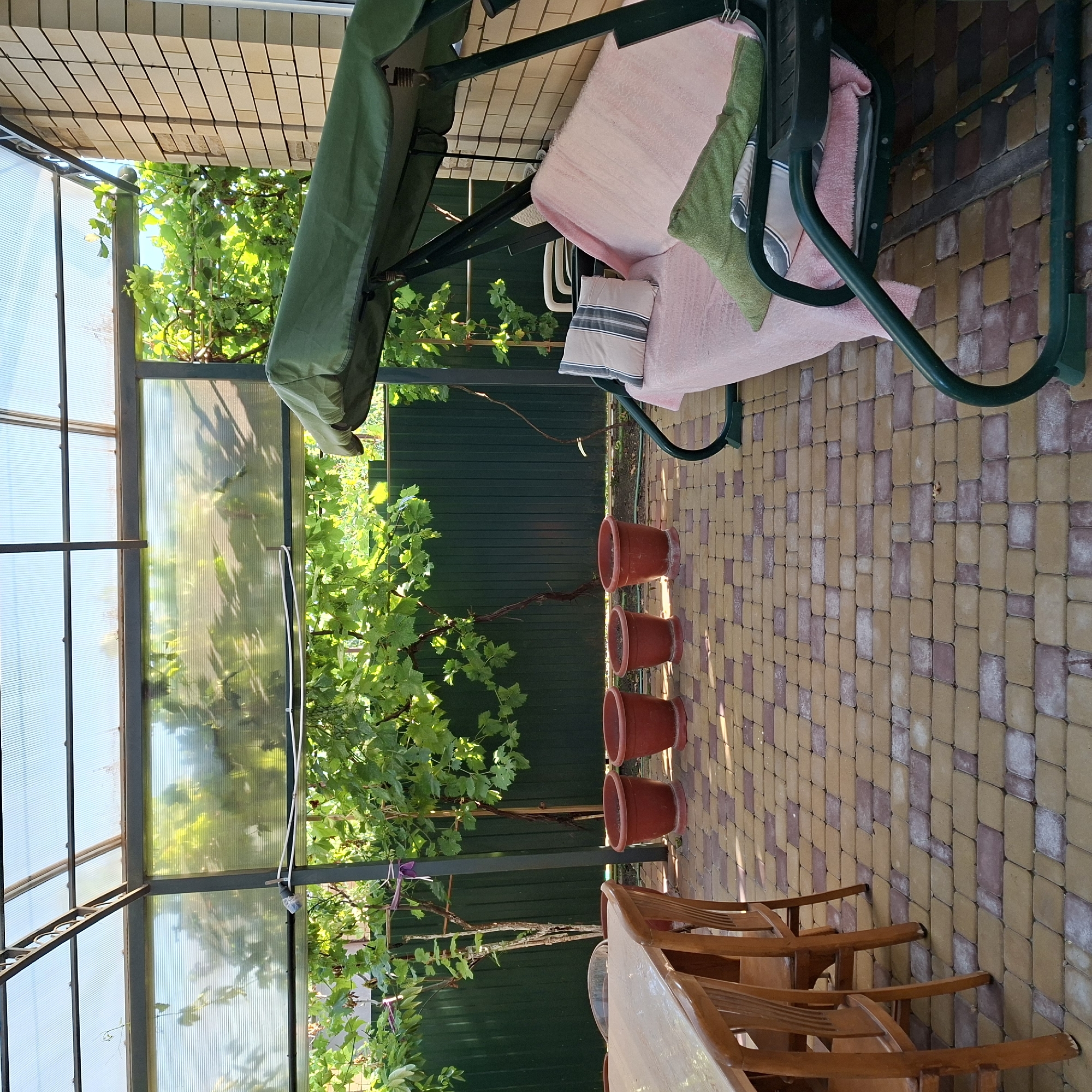 The image shows an outdoor patio area under a clear polycarbonate roof supported by a metal frame. The patio is paved with interlocking bricks in shades of yellow and burgundy. To the left, a wooden dining table with several wooden chairs is partially visible. In the background, a dark green metal fence runs along the property line, and grapevines with green leaves are trained overhead. In front of the fence, there are four terracotta-colored planters. On the right side of the patio, a green metal garden swing with a green canopy is set up. The swing is adorned with a pink blanket and two pillows, one light-colored with dark stripes and another green. The scene is well-lit, suggesting it is daytime. The location is identified as Anapskaya, Russia.