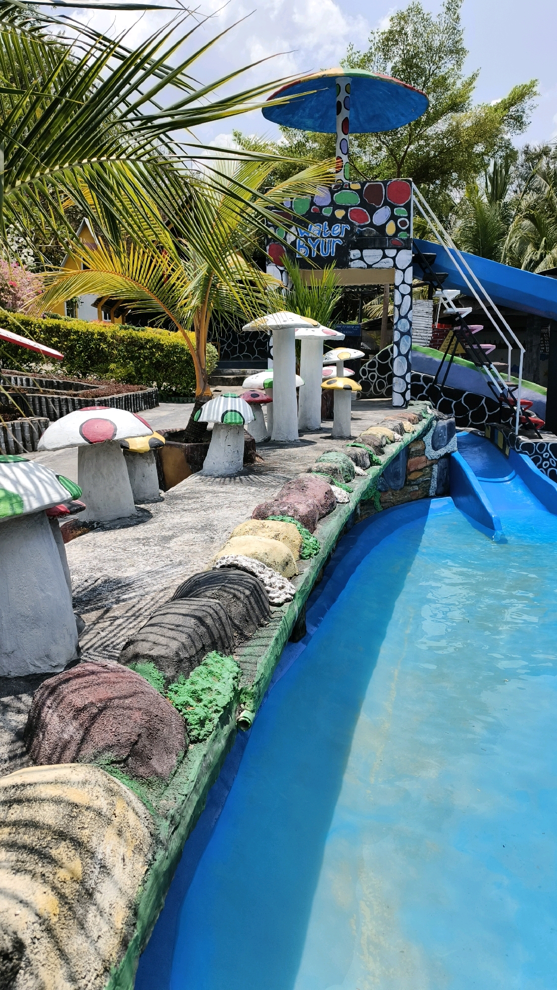 The image depicts a water park, likely located in Air Paoh, Indonesia, with a focus on a shallow, bright blue pool. The foreground features a decorative border made of what appears to be colorful, sculpted rocks and green, moss-like material, with some areas resembling tiger stripes. Along the edge of the pool, a row of mushroom-shaped structures are visible, some with red, green, or yellow polka dots on their caps.
In the background, a colorful structure with a large, multi-colored umbrella-like top stands prominently. This structure also features a blue water slide, with white steps leading up to it. The wall of this structure is adorned with various colored circles and the text "WATER BYUR". Lush green palm fronds and trees surround the water park, suggesting a tropical or subtropical environment. The sky is partly cloudy. No people are present in the image.
