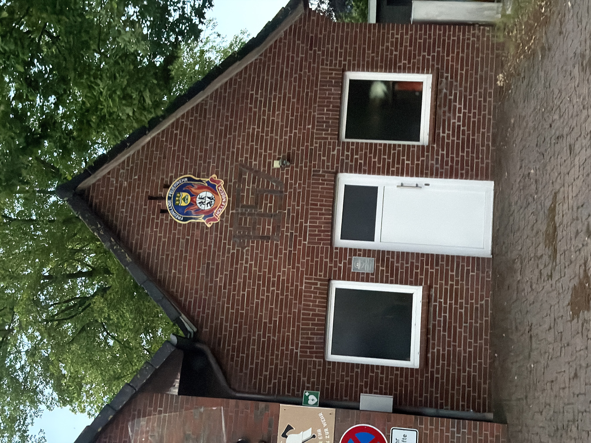 The photograph shows a brick building in Harsefeld, Germany. The building has a gabled roof, a white door, and two white-framed windows. A sign or plaque with the words "FREIWILLIGE FEUERWEHR HOLLENBECK-HARSEFELD" is prominently displayed. Below the windows and door is more signage. A cobblestone sidewalk is visible to the right of the building, and there are trees on the left.