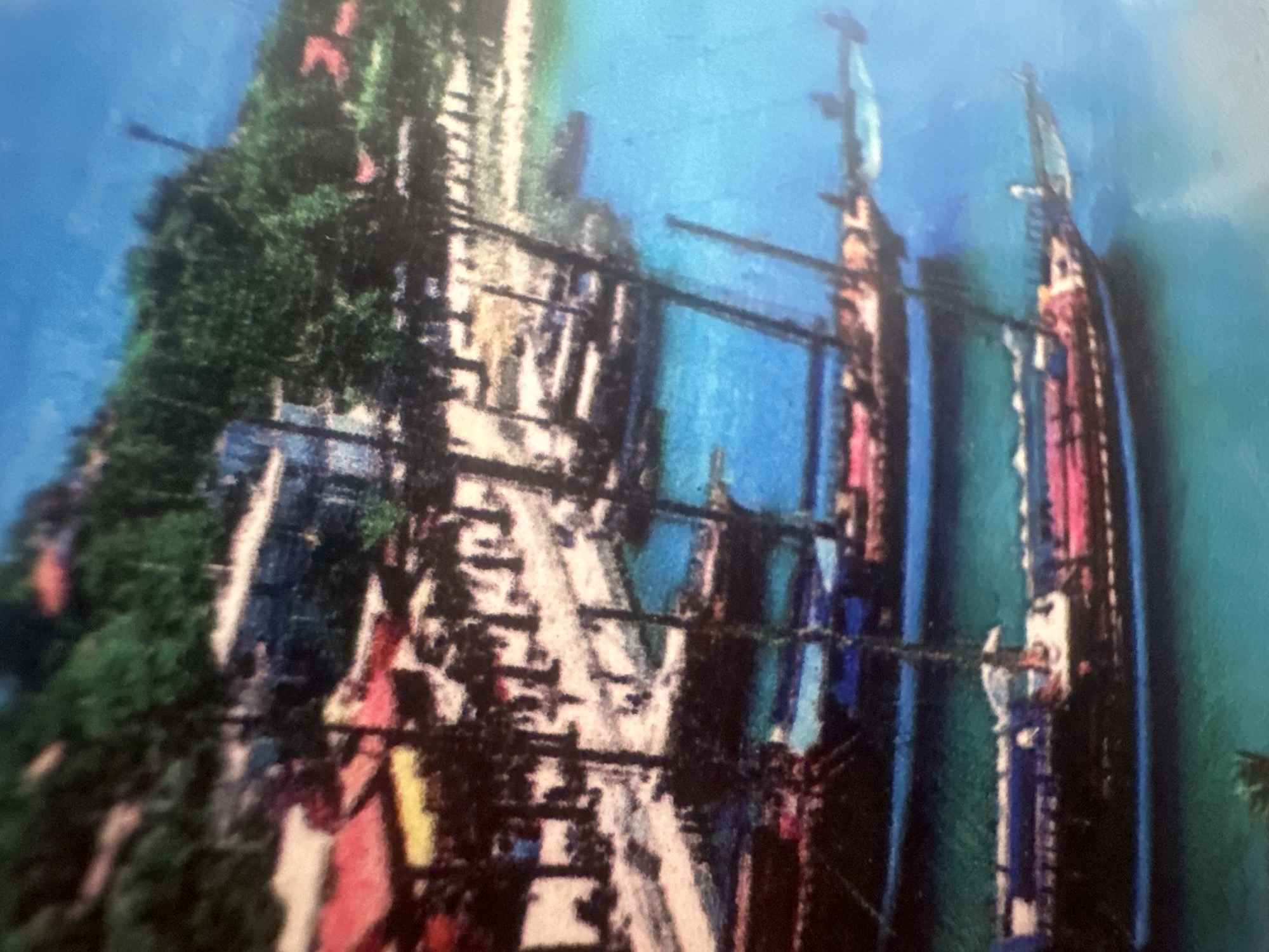 The image depicts a waterfront scene with several sailboats docked near a town or village. The water is a vibrant blue, and the boats have dark hulls and colorful decks, with masts and rigging visible. In the background, there are buildings with red and yellow roofs, nestled among lush green trees. The overall impression is one of a peaceful, tropical or Mediterranean setting, with a hint of historical charm due to the style of the boats. The location is identified as Maladzyechna, Belarus.