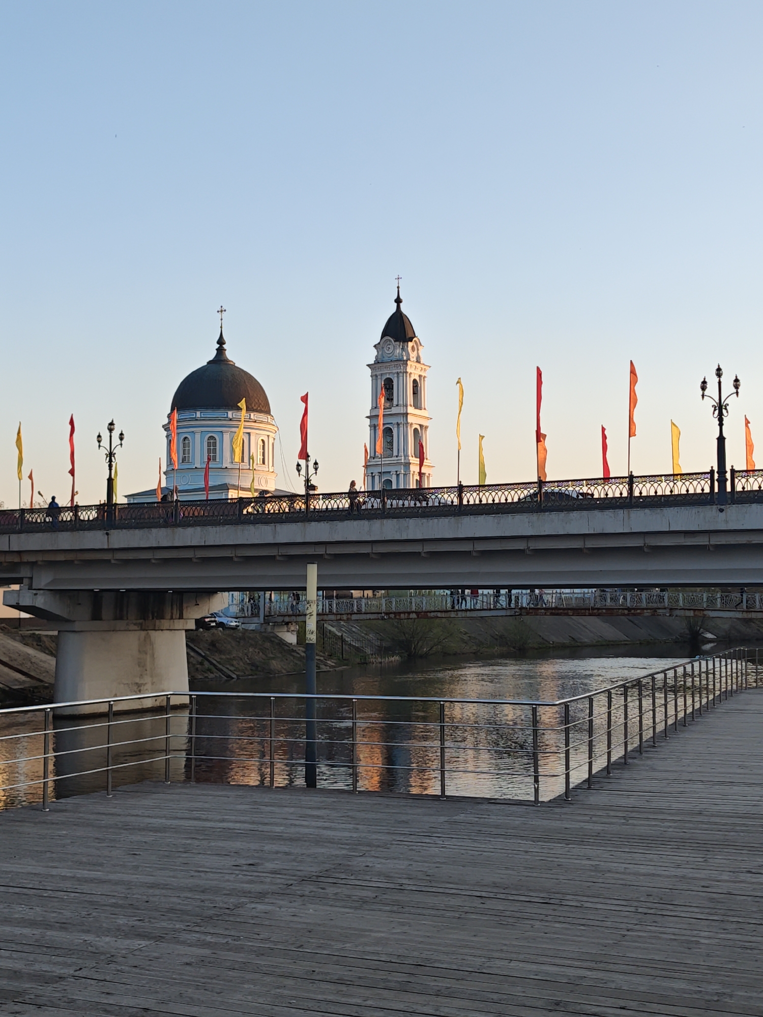 The image shows a cityscape in Noginsk, Russia, featuring the Bogoyavlensky Cathedral complex, including the cathedral itself with its distinctive dark dome and a tall bell tower. A bridge spans a river, adorned with decorative flags and lampposts, with people walking across. A wooden boardwalk extends along the riverbank in the foreground, reflecting the sunset light on the water's surface.