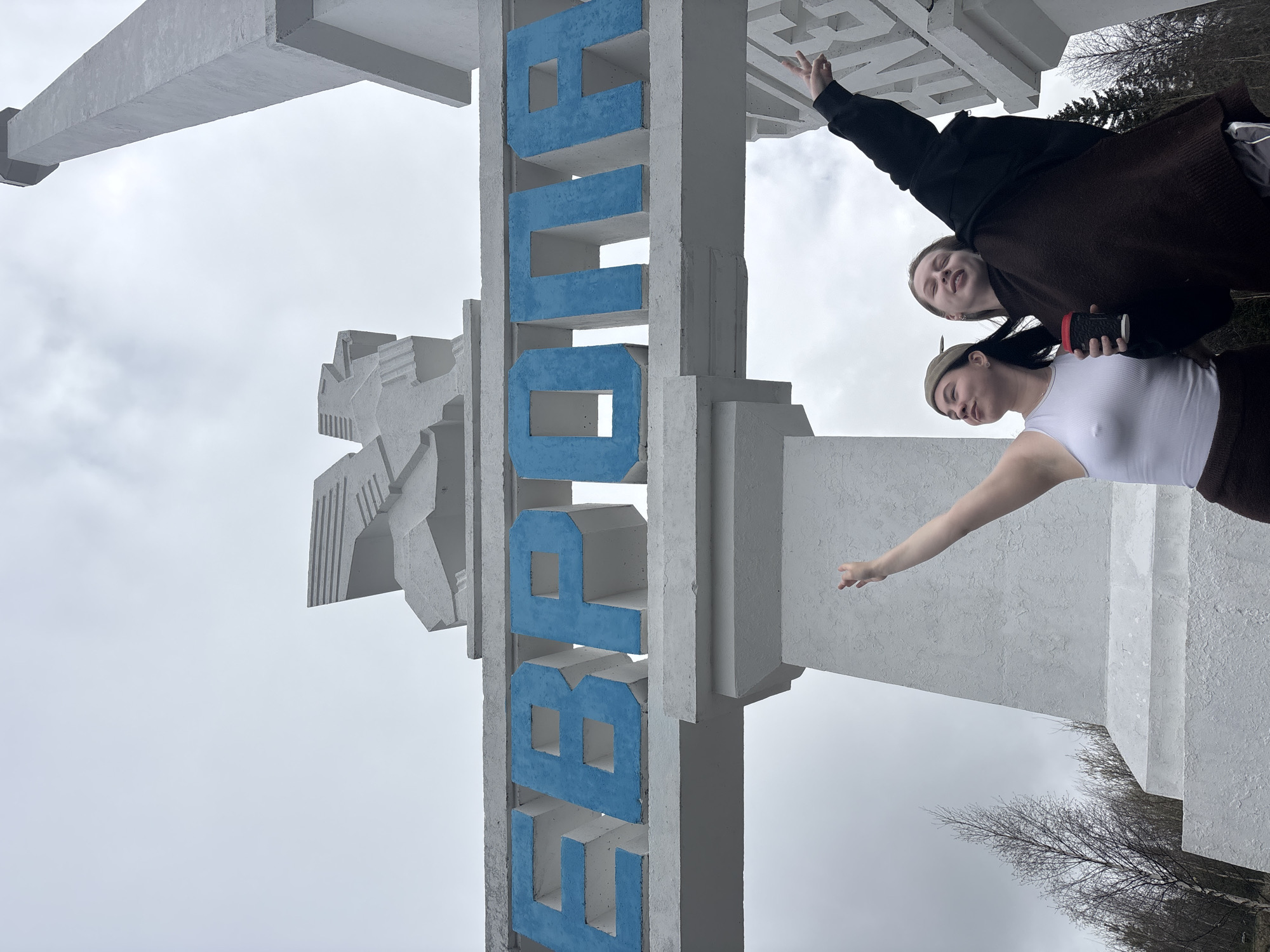 Two young women are posing in front of a large, vertical, concrete monument in the city of Promysla, Russia. The monument is predominantly white with the name of the city spelled out vertically in blue Cyrillic letters. The top woman, wearing a black jacket and brown clothing, holds up a peace sign with her right hand. The bottom woman, wearing a white top, brown clothing, and an olive-green cap, extends her left arm toward the monument. She is holding a cup in her right hand. The background shows a grey sky and bare trees.