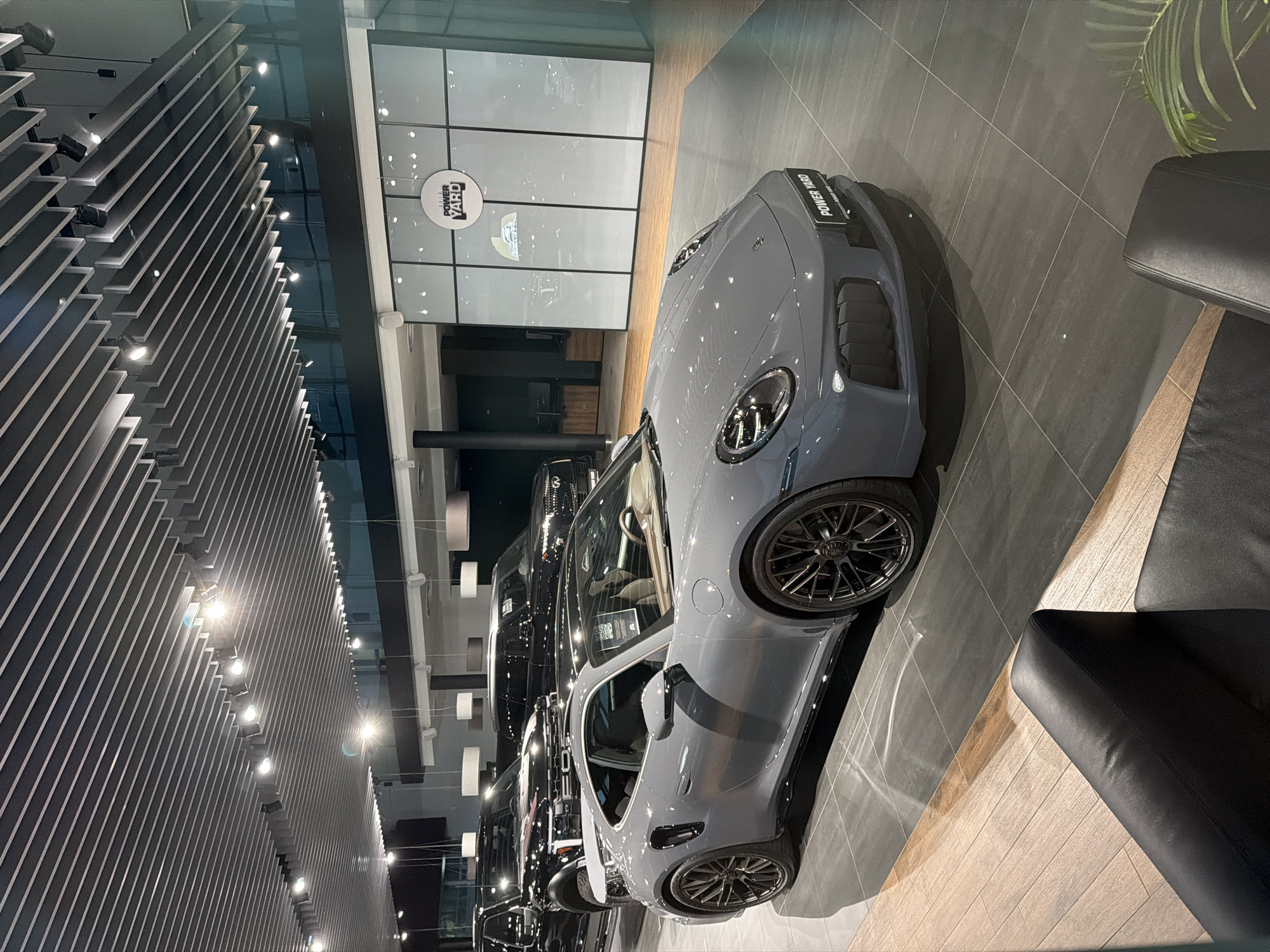 A grey Porsche 911 Turbo S is parked in a showroom with dark grey tiled flooring and wooden accent flooring. The car is positioned in the foreground, angled slightly towards the left. Behind the Porsche, two black SUVs are visible. The showroom has a modern aesthetic with a slatted ceiling and track lighting. A large glass partition with the "POWER YARD" logo is in the background. Black leather chairs are partially visible in the bottom left corner of the image. The lighting appears to be artificial and bright, casting reflections on the car and floor.