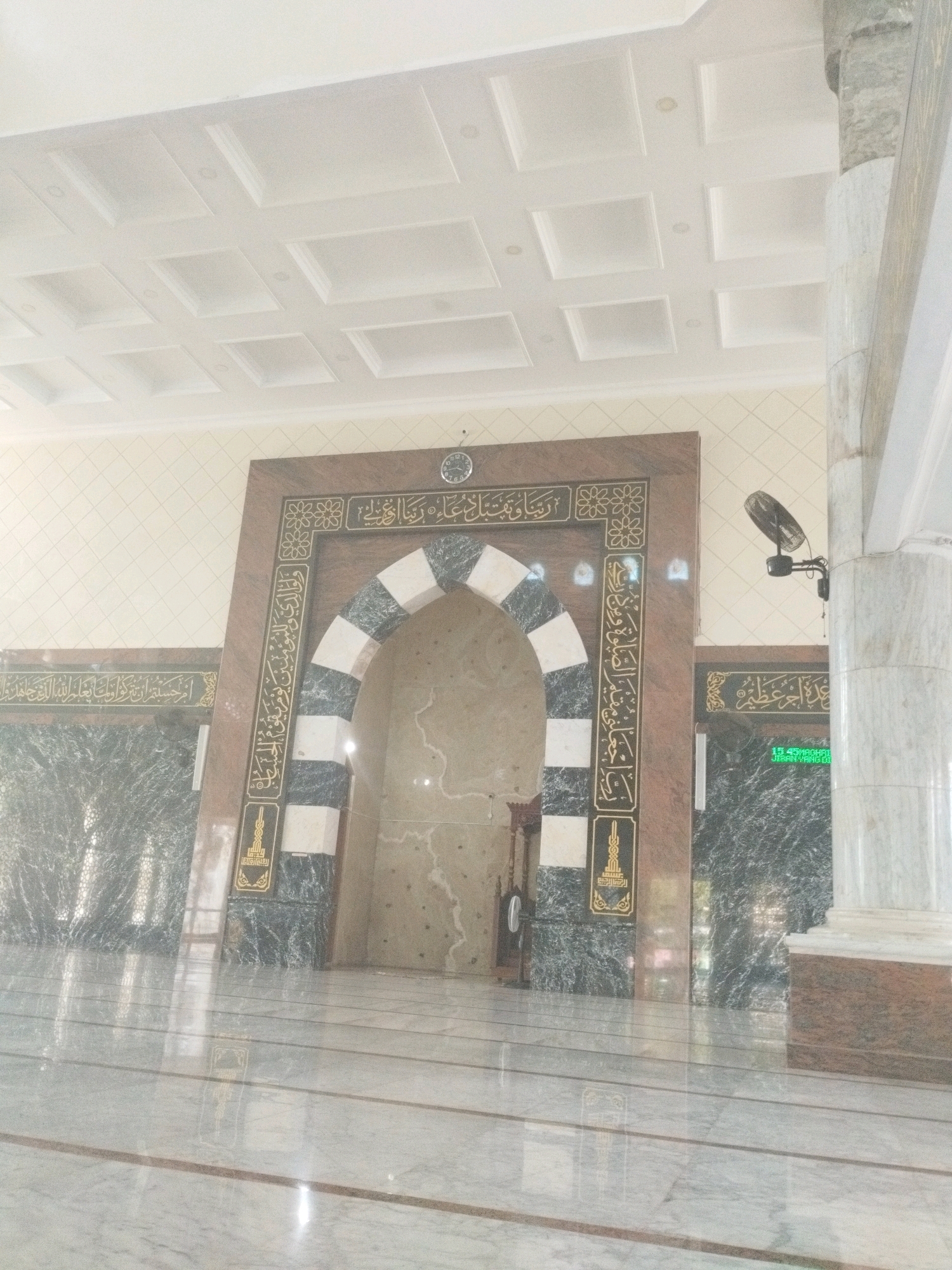 The image shows the interior of a mosque, possibly located in Tilamuta, Indonesia. A large, arched doorway with black and white marble patterns dominates the center of the frame. The doorway is adorned with ornate gold Arabic calligraphy. To the left and right of the main archway are sections of dark green marble walls also decorated with gold calligraphy. A large white marble pillar is visible on the right side of the image, supporting the ceiling. The ceiling itself is a coffered white design. The floor is polished marble, reflecting the light and architectural features of the mosque. A small electric fan is mounted on the wall near the pillar on the right, and a digital display showing "15 45 MAGHAIL" and "JIHAN YANG KU" is visible on the green marble wall. The scene is well-lit, with soft light illuminating the space. There are no people visible in the image.