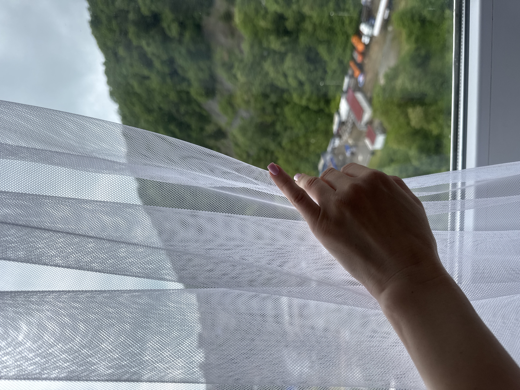 A person's hand is opening a window blind to reveal an outdoor scene. The view outside the window appears to be of a hillside densely covered in trees, with a row of buildings or structures visible in the distance. Based on the landscape and background details, it is located in the city of Sochi, Russia.