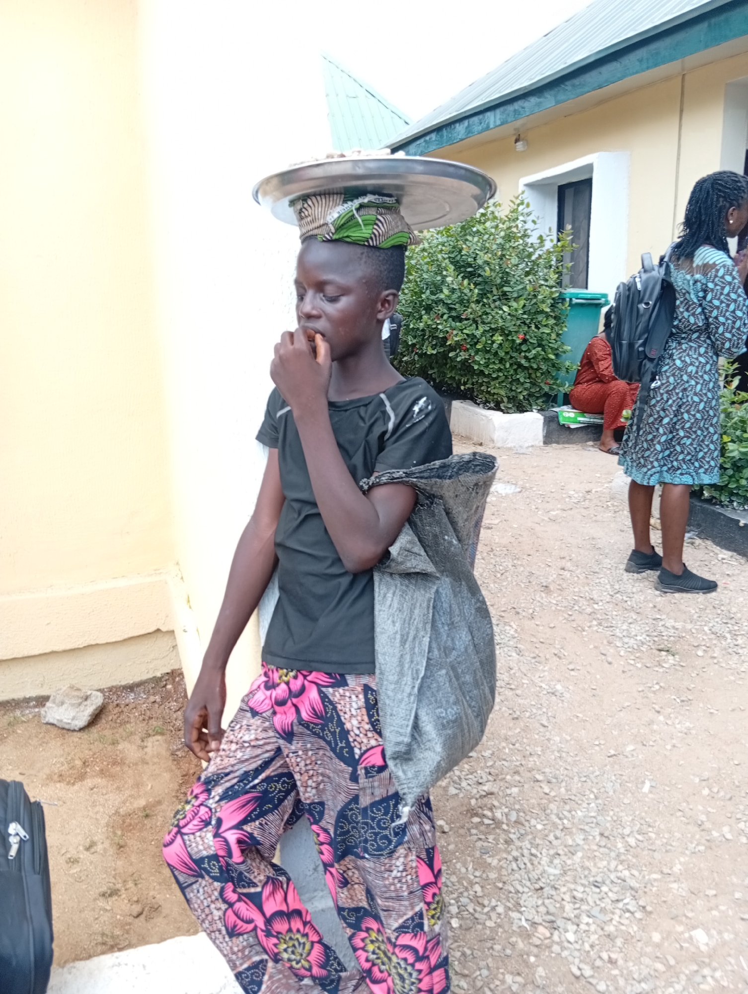 A young person is standing outdoors in the city of Jalingo, Nigeria, with a tray of food balanced on their head. The individual is wearing a black t-shirt and brightly patterned pants with pink and blue floral designs. They have a grey fabric bag slung over their right shoulder. The background features a light-colored building with a green trim and a dense green bush. Another person, wearing a blue and black patterned dress, is standing to the right, looking away from the camera and carrying a black backpack. Further in the background, a person in a red outfit is seated. The ground is covered in gravel and sand, with a black suitcase visible to the left of the main subject. The overall impression is of a street scene with people engaged in daily activities.