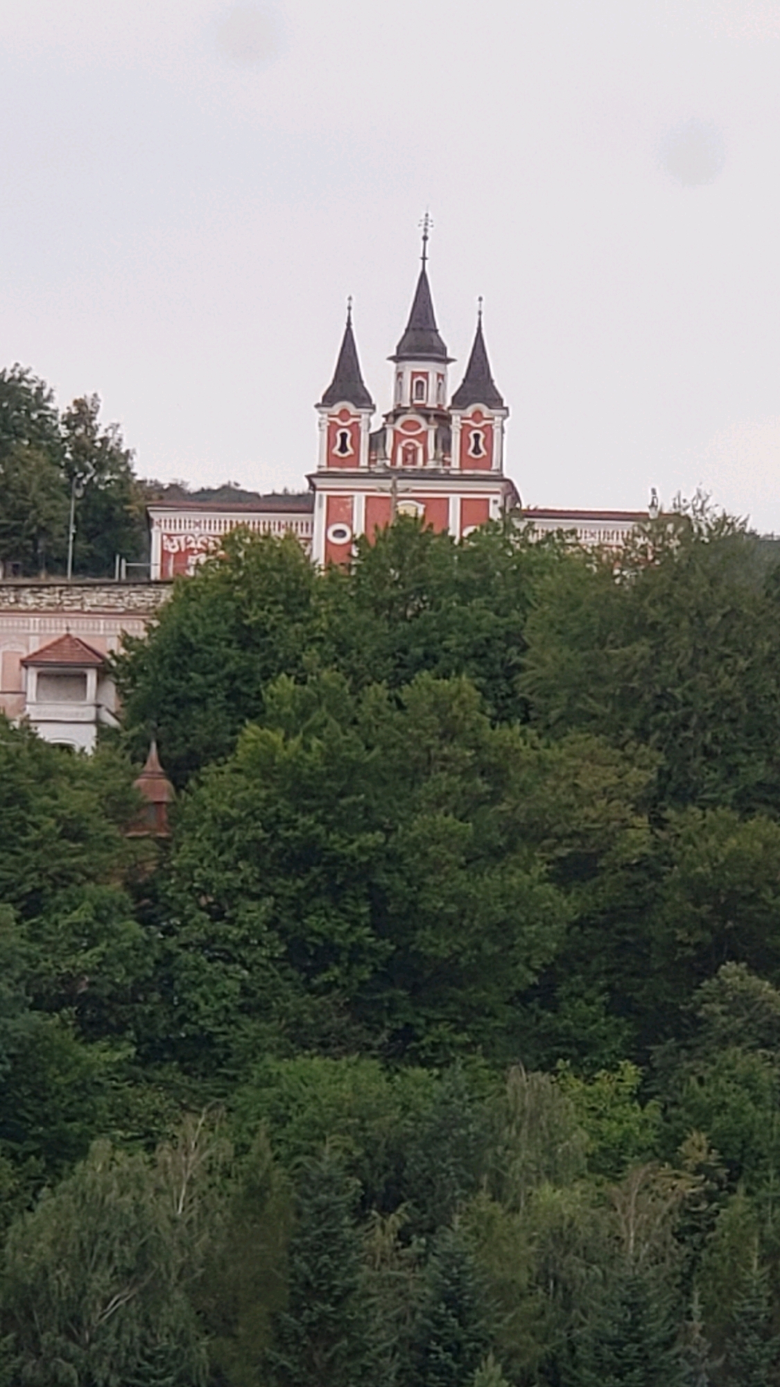 The image shows a view of the Church of the Nativity of the Virgin Mary in Prešov, Slovakia, situated atop a hill and surrounded by lush green trees. The church features a prominent red facade with white trim and three dark, pointed spires. The central spire is taller and topped with a cross, while the two flanking spires have decorative elements. Below the main body of the church, a section of white building with a small, dark-roofed structure can be seen on the left. The foreground is filled with a dense canopy of various trees, with the overcast sky forming the backdrop.