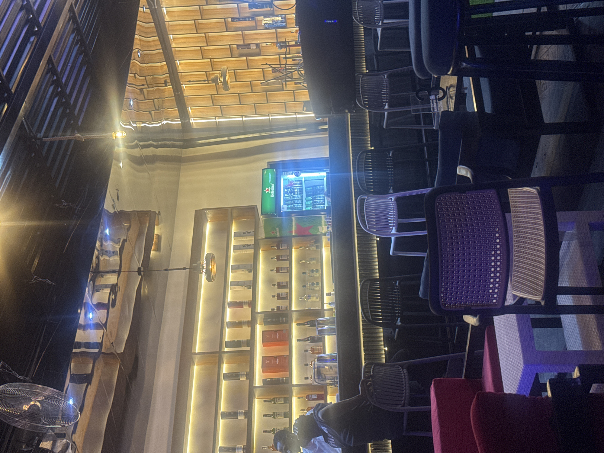 The image depicts the interior of a modern bar and lounge, likely located in Abuja, Nigeria. The scene is dimly lit, with warm ambient lighting emanating from shelves stocked with bottles of liquor and from decorative light fixtures. The bar area is prominent, with a long counter and an array of stools. A refrigerator displaying green bottles, possibly beer, is visible to the right of the bar. In the foreground, several chairs and small tables are arranged, suggesting a seating area. A ceiling fan is visible in the upper left corner, contributing to the ambiance. The decor features a mix of wooden elements and metallic finishes, creating a sophisticated and contemporary atmosphere.