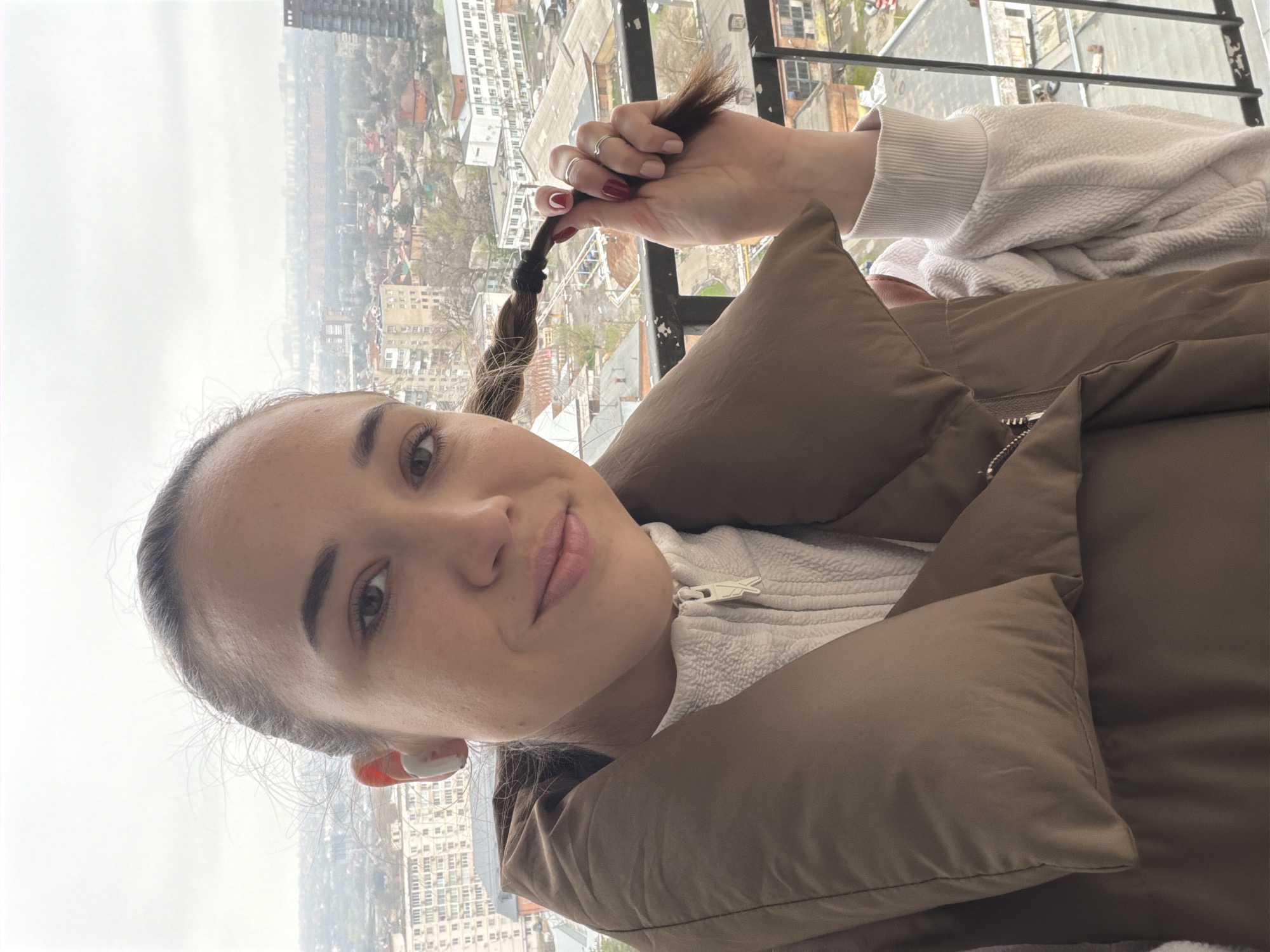 A selfie image shows a woman with her hair in a braid, standing on a balcony overlooking the city of Rostov-on-Don, Russia. She is wearing a brown puffer jacket and a light-colored sweater. Her fingernails are painted with dark red nail polish, and she has white earbuds in her left ear. She is holding the end of her braid in her hand. In the background, buildings and trees are visible, with a gray overcast sky.