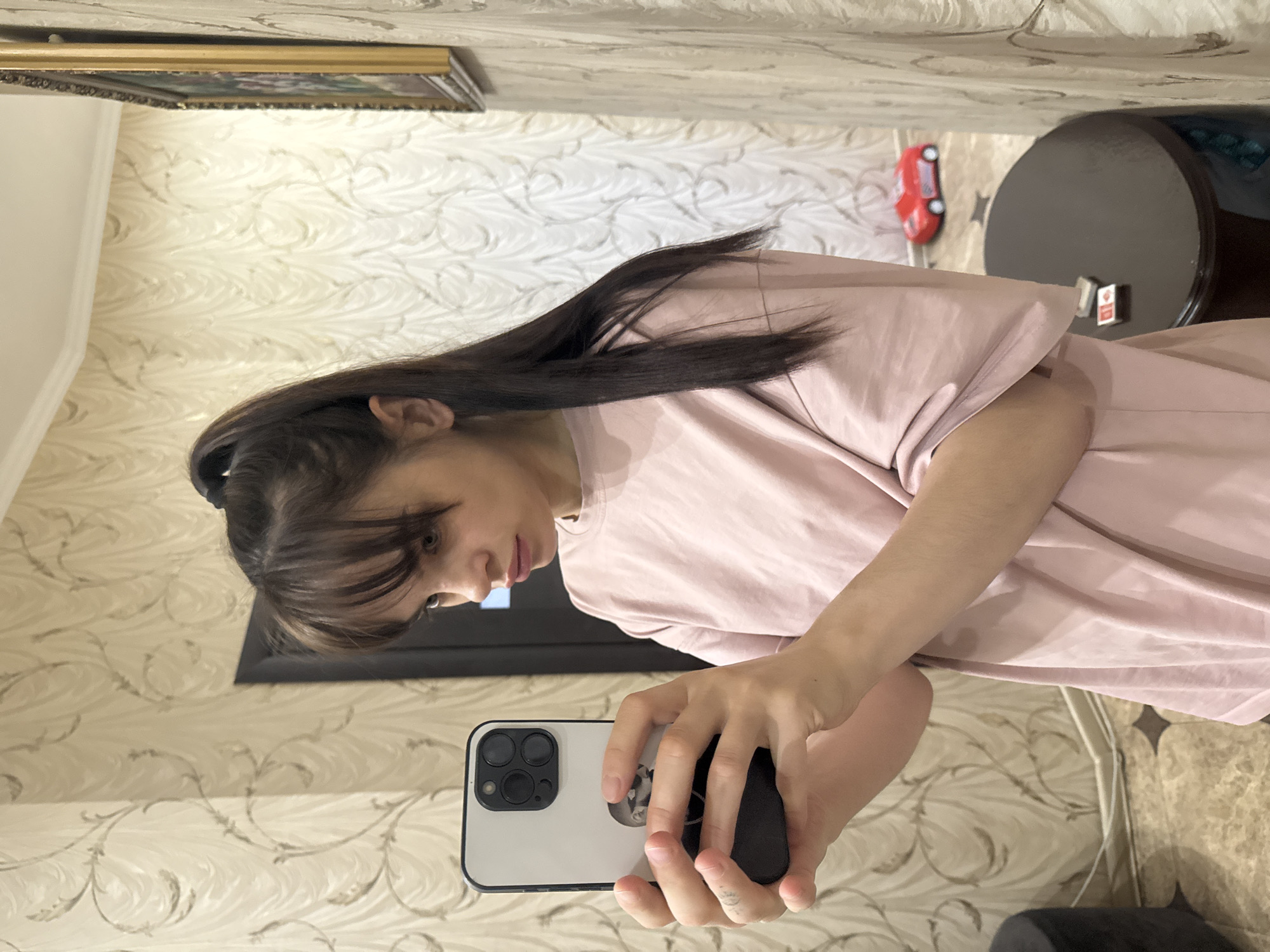 A woman is taking a selfie with a smartphone in a room with floral patterned wallpaper. The room contains a TV, a round table, and framed artwork. A toy car is on the table. It is not possible to confirm if this image was taken in the city of Rostov-on-Don, Russia.