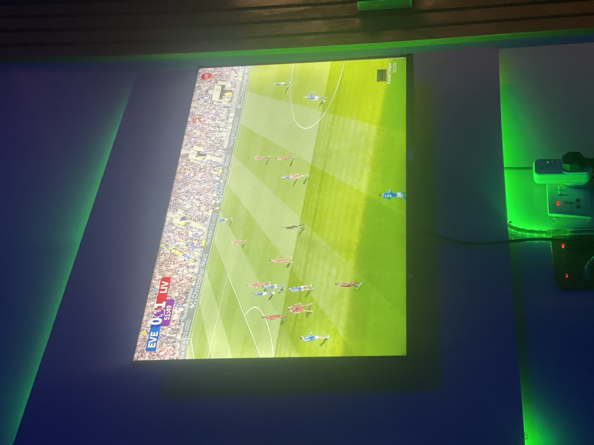 The image shows a television screen displaying a live soccer match. The score is Everton 0, Liverpool 1, with the time at 51:49. The Premier League logo indicates the 2025/26 season. The screen also displays advertisements for "The NEW C12 LOCO" and "Christopher Ward." On the right of the screen, a wall socket with a plug is visible. The environment around the television is dark, with green lighting. The location is unknown.