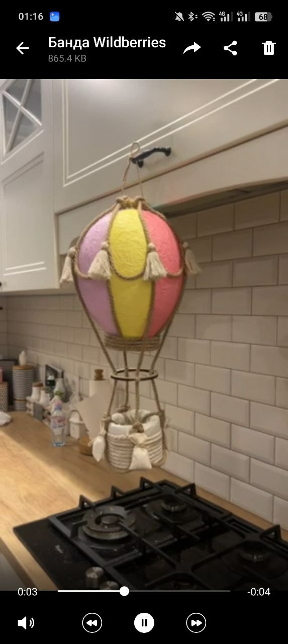 The image shows a hot air balloon decoration hanging in a kitchen. The balloon is pink and yellow with rope detailing and tassels. It hangs above a kitchen countertop with a black stovetop. The background includes white cabinets and a white tile backsplash. The image appears to be a screenshot from a video, as indicated by the play controls at the bottom. While the image lacks specific details confirming the location, the presence of Russian text at the top ("Банда Wildberries") suggests a region where Russian is spoken, potentially Minsk, Belarus. However, this is speculative without further evidence.