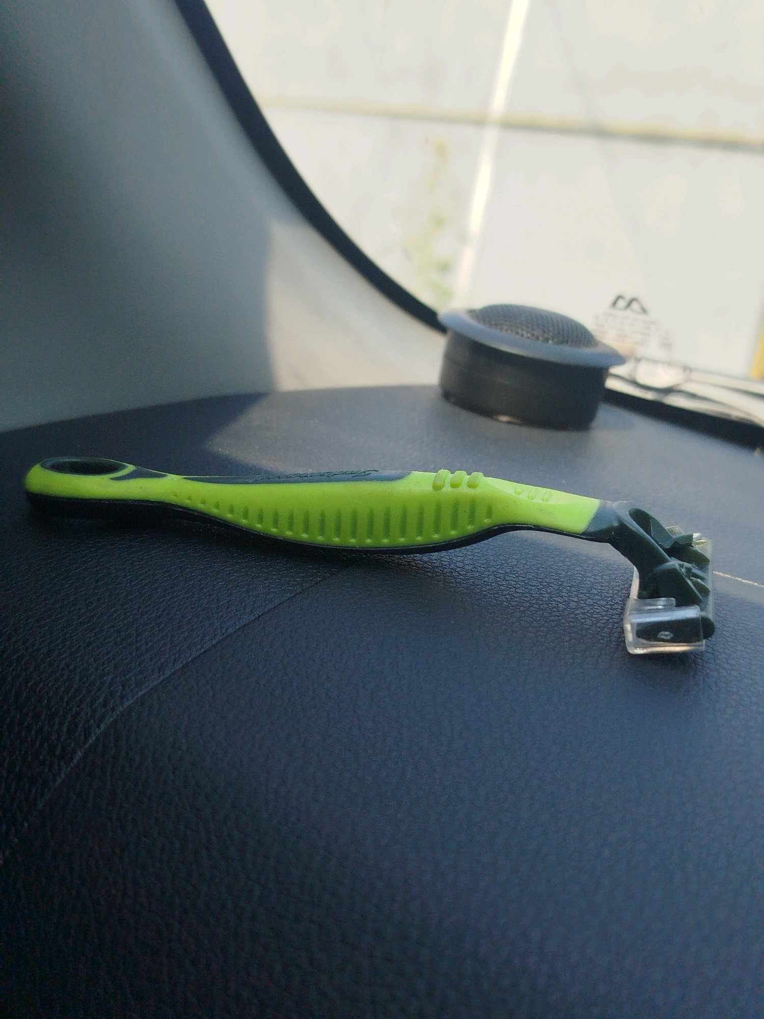 A green and black razor lies on a dark, textured surface, possibly a car dashboard. The razor has a textured grip and a clear plastic head with visible blades. In the background, a black circular object with a mesh surface is visible, along with some blurry white and yellow shapes suggesting a sunlit environment. The location of Cagakagung, Indonesia is not depicted in the image.