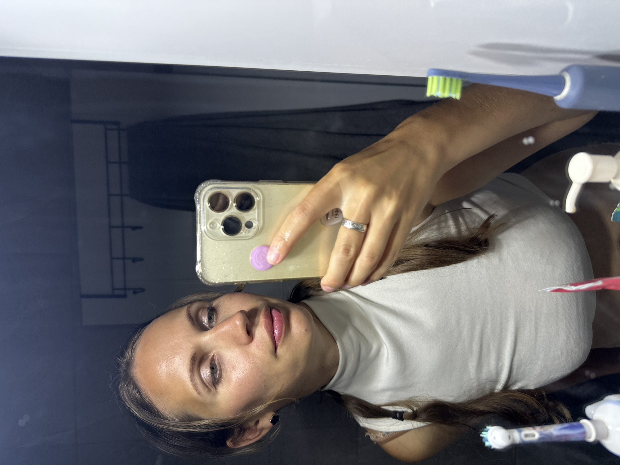 A woman is taking a selfie in a bathroom. She is holding a smartphone with a clear case and a purple circular object on the back. She is wearing a white top. In the reflection of the mirror, there are toothbrushes and other bathroom items. The location is a bathroom, details that would identify the specific location as Fethiye, Turkey are not visible in the image.