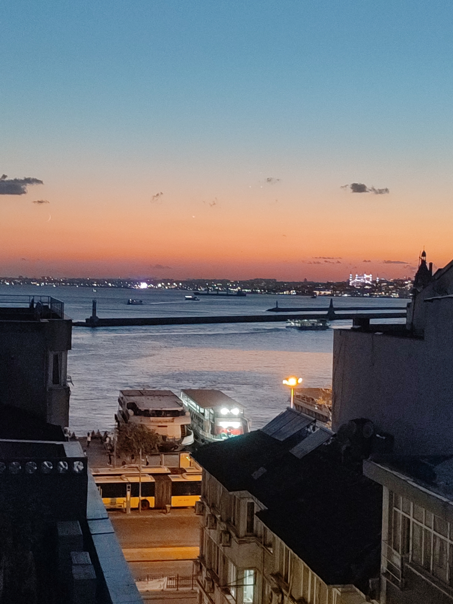 The image captures a sunset view over the Bosphorus Strait, likely from Kadıköy, Turkey, given the silhouette of the Hagia Sophia and Blue Mosque in the distance on the European side. The sky transitions from a deep blue at the top to vibrant orange and pink hues near the horizon. Several boats, including passenger ferries, navigate the water. A lighthouse stands on a breakwater extending into the strait. In the foreground, buildings are visible, with what appears to be a street or waterfront promenade lined with buses and some pedestrians. The overall scene is one of urban life by the water during the transition from day to night.