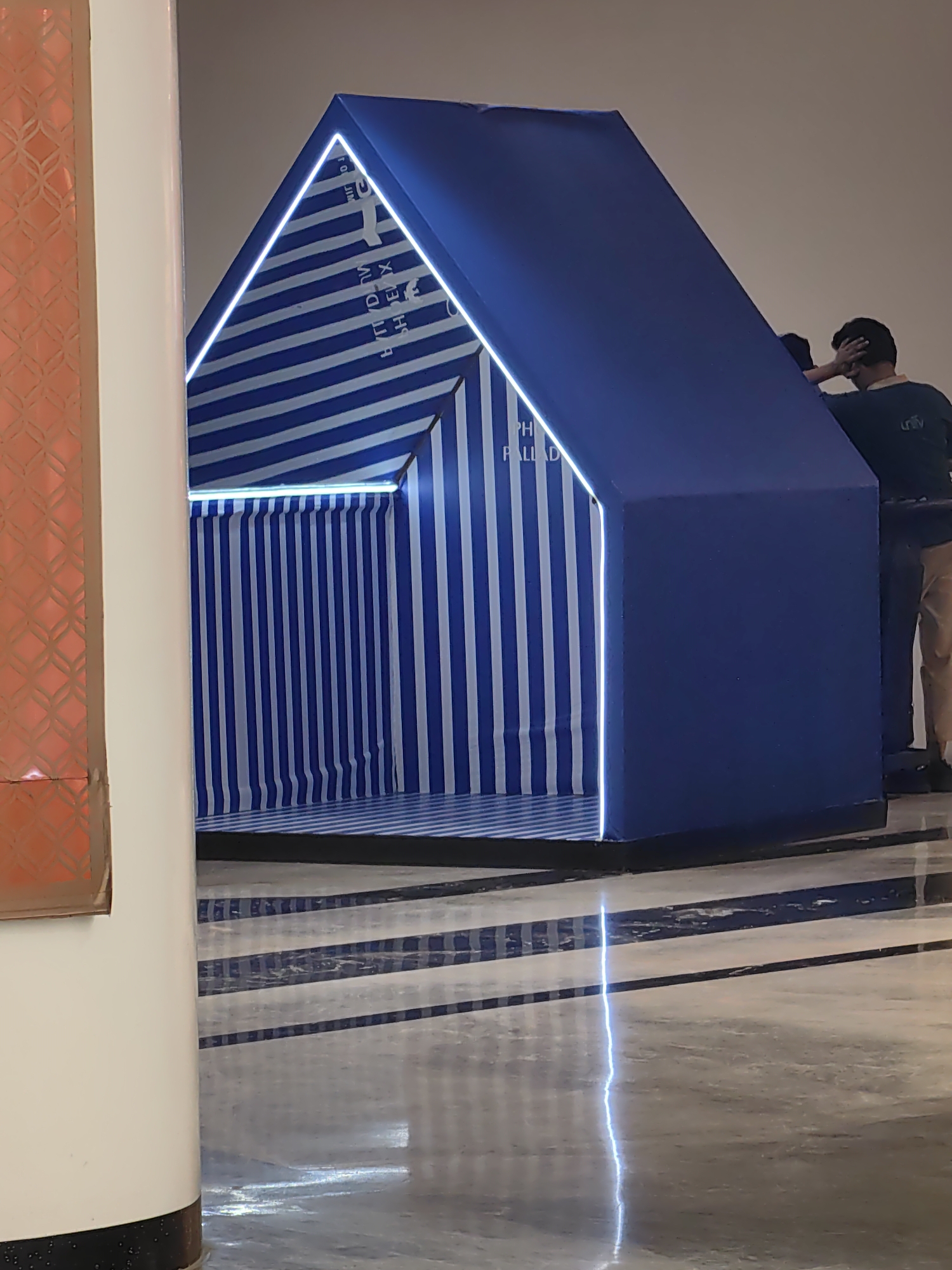 An indoor, overhead shot shows a stylized, blue, prism-shaped structure with illuminated white edges, situated on a reflective floor patterned with dark blue and white stripes. The structure's interior is visible and also features blue and white stripes. Text is visible on the interior of the structure, which appears to be upside down and reversed. To the right of the structure, two individuals are partially visible. One person, wearing a dark blue shirt and khaki pants, is standing with their hand on their head, looking towards the structure. Another person, wearing a dark top, is mostly obscured by the structure. A white column with a patterned, orange design is visible on the left side of the frame. The background appears to be a plain, light-colored wall. The scene is likely set in a public indoor space, such as a mall or exhibition hall, possibly in Mumbai, India, given the presence of the Palladium logo on the structure's interior.
