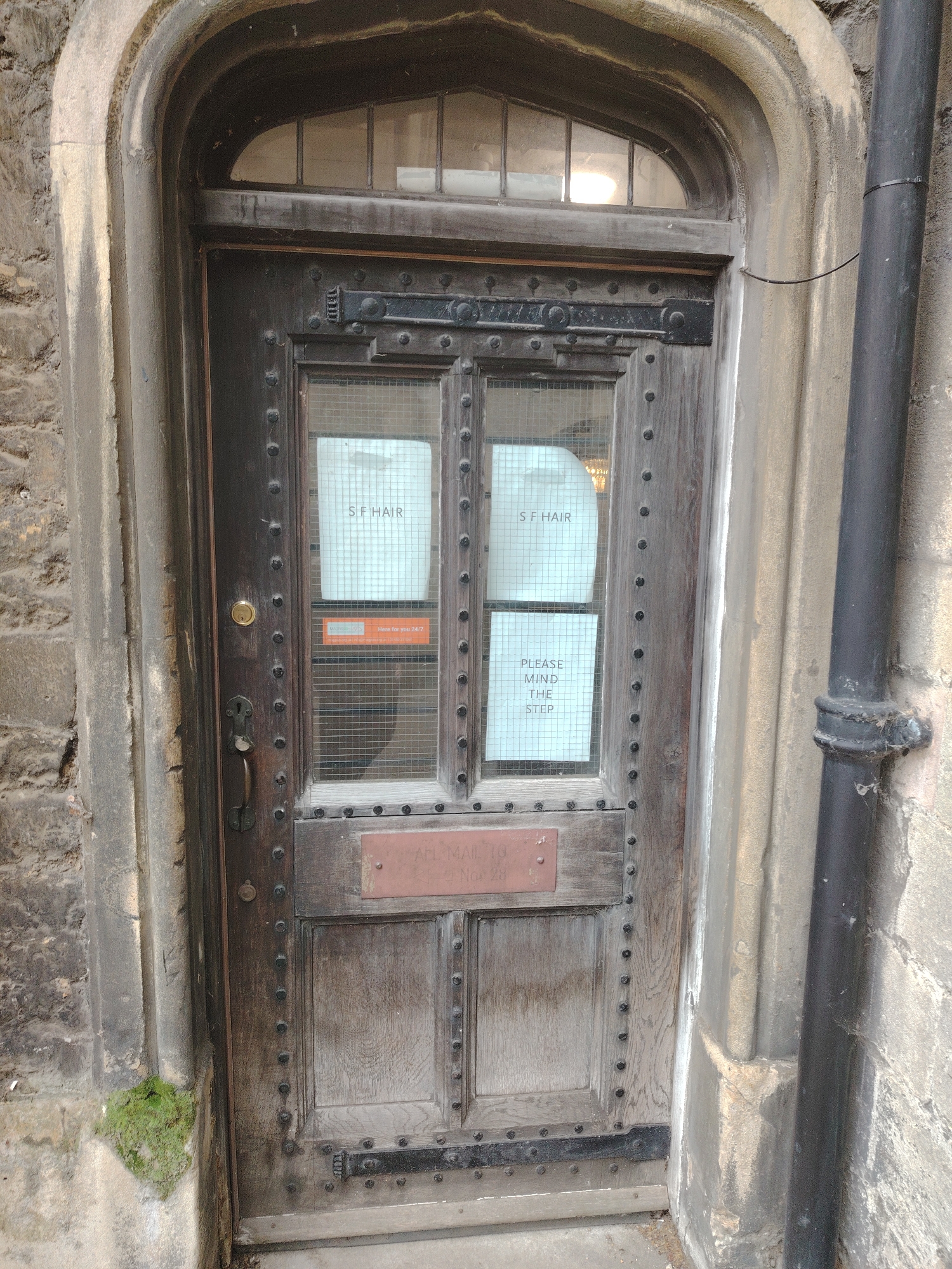 The image depicts a weathered, dark wooden door recessed within an arched stone facade in Peterborough, United Kingdom. The door features an arched multipane transom window at the top and two central glass panels, each covered with a metal grid. White paper signs are affixed to the interior of these panels, reading "S.F HAIR" on both upper panes and "PLEASE MIND THE STEP" on the lower right pane. A small orange sticker with text including "MAZARS" and "here for you 24/7" is partially visible below the left "S.F HAIR" sign.
The door is adorned with numerous black, round studs arranged vertically and horizontally. It is equipped with an ornate, dark metal handle, a circular keyhole above the handle, and a reddish-brown rectangular metal plate (likely a mail slot cover) below the glass panels. The lower section of the door consists of paneled wood, also decorated with studs.
To the left of the door, a rough-hewn stone wall is visible, with a small patch of green moss growing at its base near the doorframe. To the right, a black cylindrical downspout extends vertically against the stone wall, with a black cable secured to it by a zip tie. The stone archway and surrounding walls exhibit significant weathering and discoloration. The ground directly in front of the door appears to be a light-colored, worn step or pavement. No individuals are present, and no actions or direct interactions are depicted in the scene.