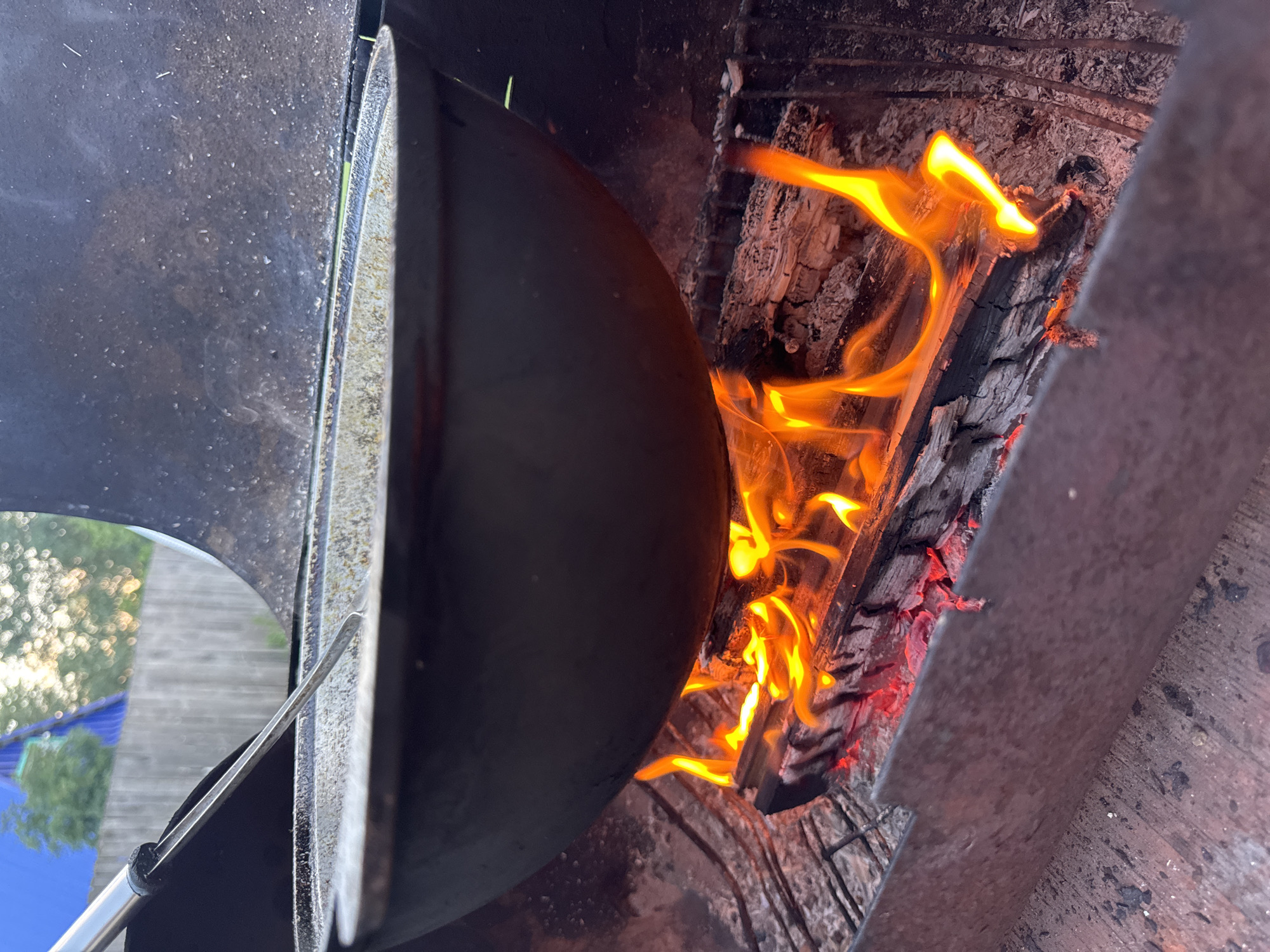 A cast-iron pot is suspended over a fire in an outdoor setting, likely a backyard in Chaikovsky, Russia, indicated by the presence of a wooden fence and residential buildings in the background. The fire, fueled by logs, produces vibrant orange and yellow flames, casting a warm glow on the surrounding area. Inside the pot, a portion of what appears to be a meal is visible, with a metal utensil resting on the rim, suggesting preparation or cooking is in progress. The overall scene captures a moment of outdoor cooking, emphasizing the rustic and primal nature of preparing food over an open flame.