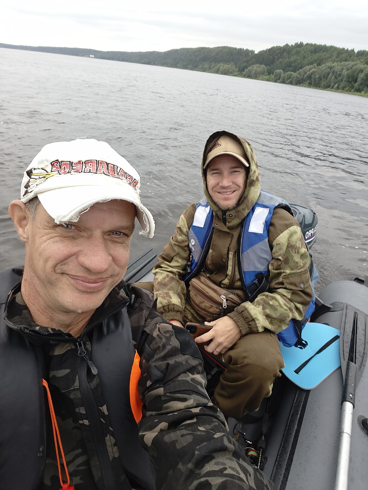 Two men are in an inflatable boat on a river. The man in the foreground is wearing a white baseball cap with red and black lettering and a black and camouflage jacket with an orange life vest underneath. He is smiling at the camera. The man in the background is wearing a camouflage hooded jacket and a blue and grey life vest. He is also smiling. The boat has a small outboard motor. An oar is visible next to the man in the background. There is also a blue seat cushion and a fishing rod with a reel. The river is wide and calm, with a tree-lined bank in the distance. The sky is overcast, suggesting it is daytime, possibly morning or afternoon. The overall mood is relaxed and friendly. The lettering on the man's cap is partially visible but unreadable.