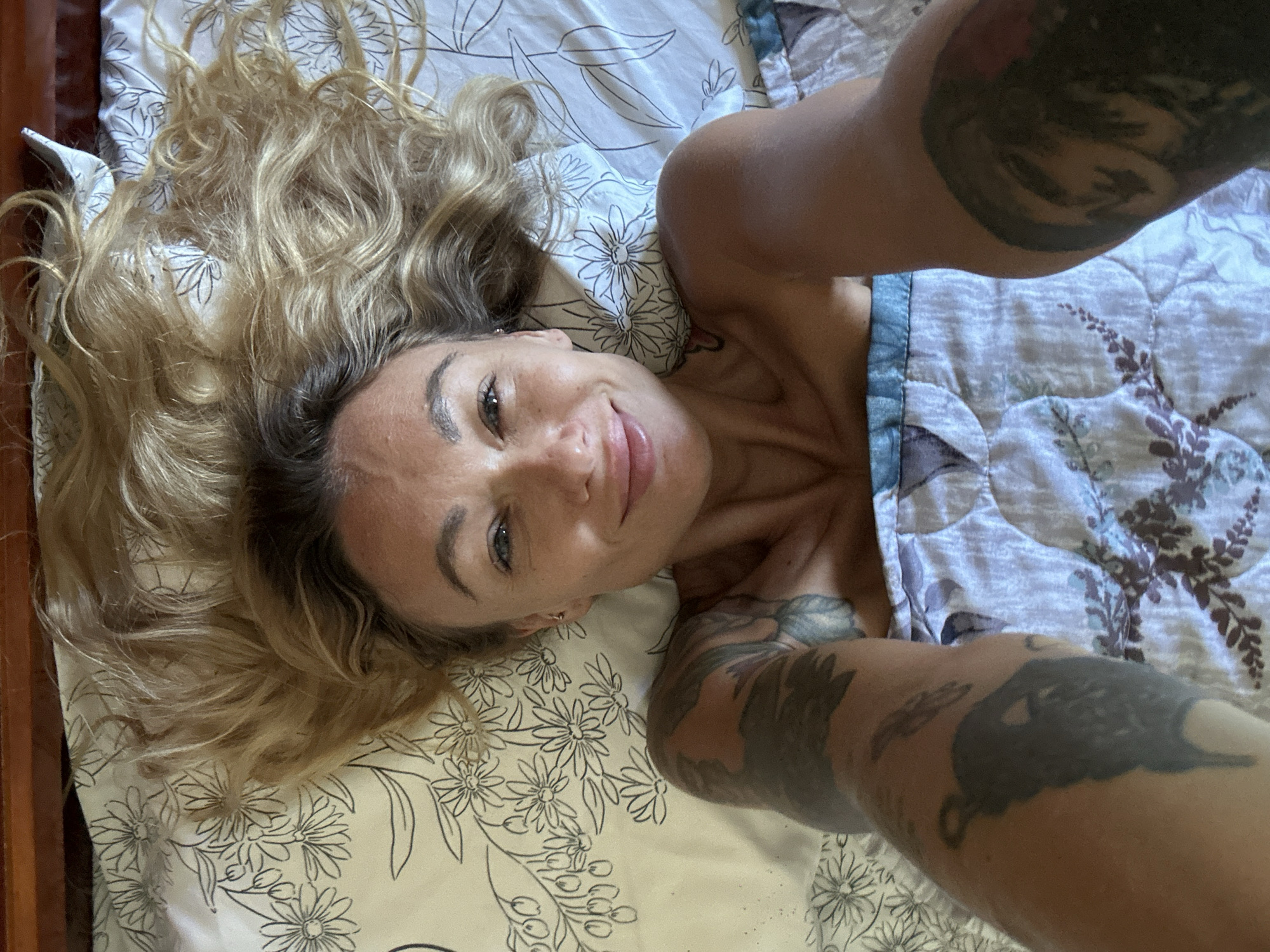 A woman with blonde, wavy hair is lying in bed, looking up at the camera and smiling. She is wearing a blue and white patterned blanket. Her left arm, which is tattooed, is extended towards the camera. The bed is covered with a white floral patterned sheet and a quilted blanket with a floral pattern in purple and blue. The background is a wooden headboard. The image does not provide any information about the location in the city of Mũi Né, Vietnam.