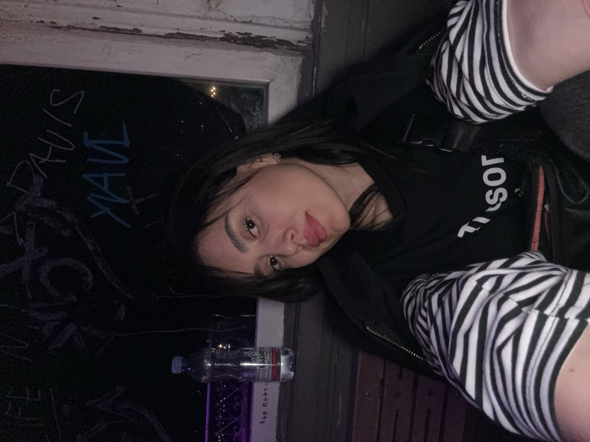 A young woman is depicted in a rotated, close-up portrait, appearing to be lying down or leaning against a surface. She has long, dark hair, dark eyebrows, and a playful expression, looking directly at the camera with her tongue slightly extended. She is wearing a black long-sleeved top or jacket with white, upside-down text that reads "SOR" on the chest. Her arms are covered in black and white striped sleeves or a striped undergarment. A small, shiny earring is visible on her left ear.
The setting is dark, suggesting nighttime or low light conditions. Behind her is a dark wall or surface adorned with white and light blue graffiti; discernible text includes "PAUIS" and "YAV." Some indistinct purple scribbles are also present lower down. To her left, a worn window frame with peeling white paint is visible, and through the window, some distant lights can be seen.
Resting on a ledge next to her is a clear plastic water bottle with a red and white label. The label prominently displays the Georgian text "ნაბეღლავი" (Nabeglavi), indicating it is a bottle of Georgian mineral water, and also states "0.5L." The overall mood is casual.