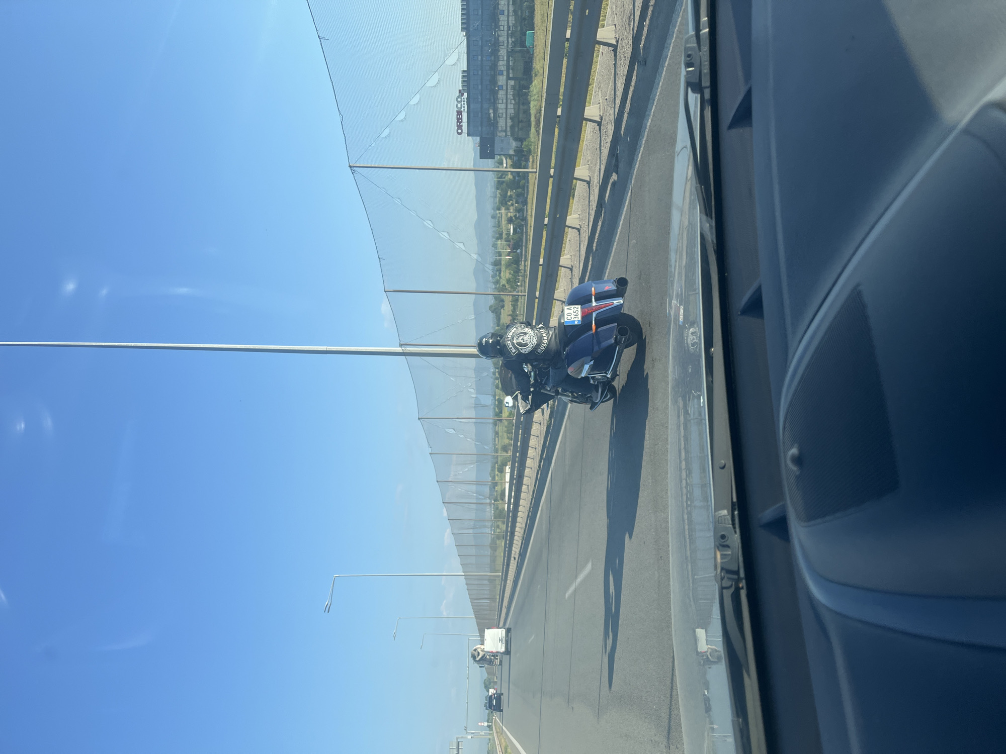 An outdoor, eye-level, daytime, long shot captures a view from the passenger seat of a vehicle traveling on a highway. The road is paved and appears to be a multi-lane highway with a concrete barrier separating the lanes. A motorcyclist in a black helmet and black leather jacket rides a blue motorcycle in the right lane. The motorcycle has saddlebags and a license plate that reads "CO A 3652". In the background, there are distant mountains and a building with the word "ORBICOM" visible on its side. A tall light pole stands to the left of the frame, and a mesh fence runs along the right side of the highway. The sky is clear and blue.