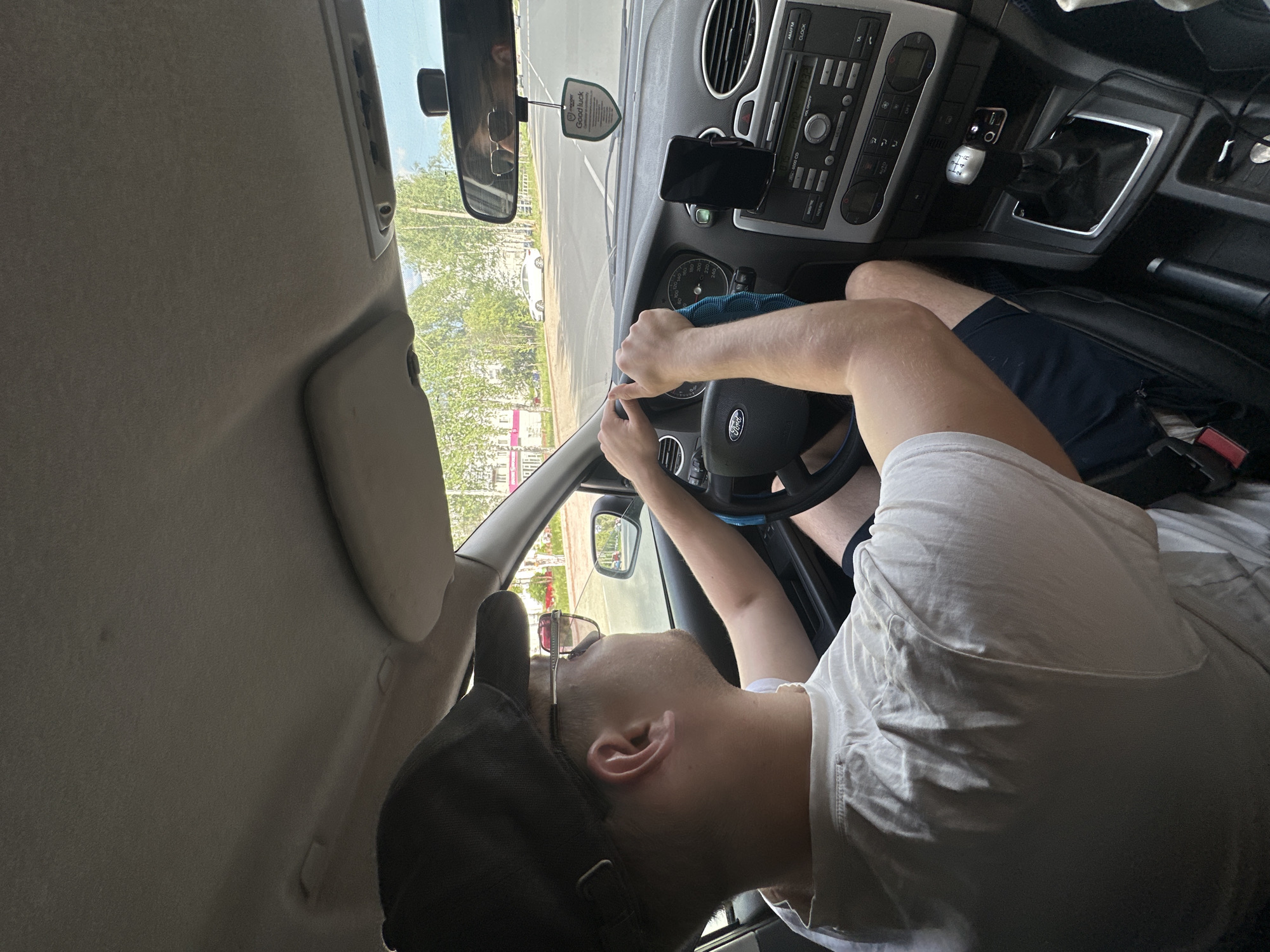 The image shows a man sitting in the driver's seat of a car. He is wearing a white t-shirt, dark shorts, and a black baseball cap. The car's dashboard is visible, featuring a Ford steering wheel and a manual transmission gear shift. A mobile phone is mounted on the dashboard. The rearview mirror displays a reflection of the street outside, including a building with a pink sign. Based on the sign's text "МАГАЗИН ЧАЙКОВСКИЙ", it is likely the photograph was taken in Chaikovsky, Russia. The man is gripping the steering wheel with his hands, with a blue cloth in his left hand.
