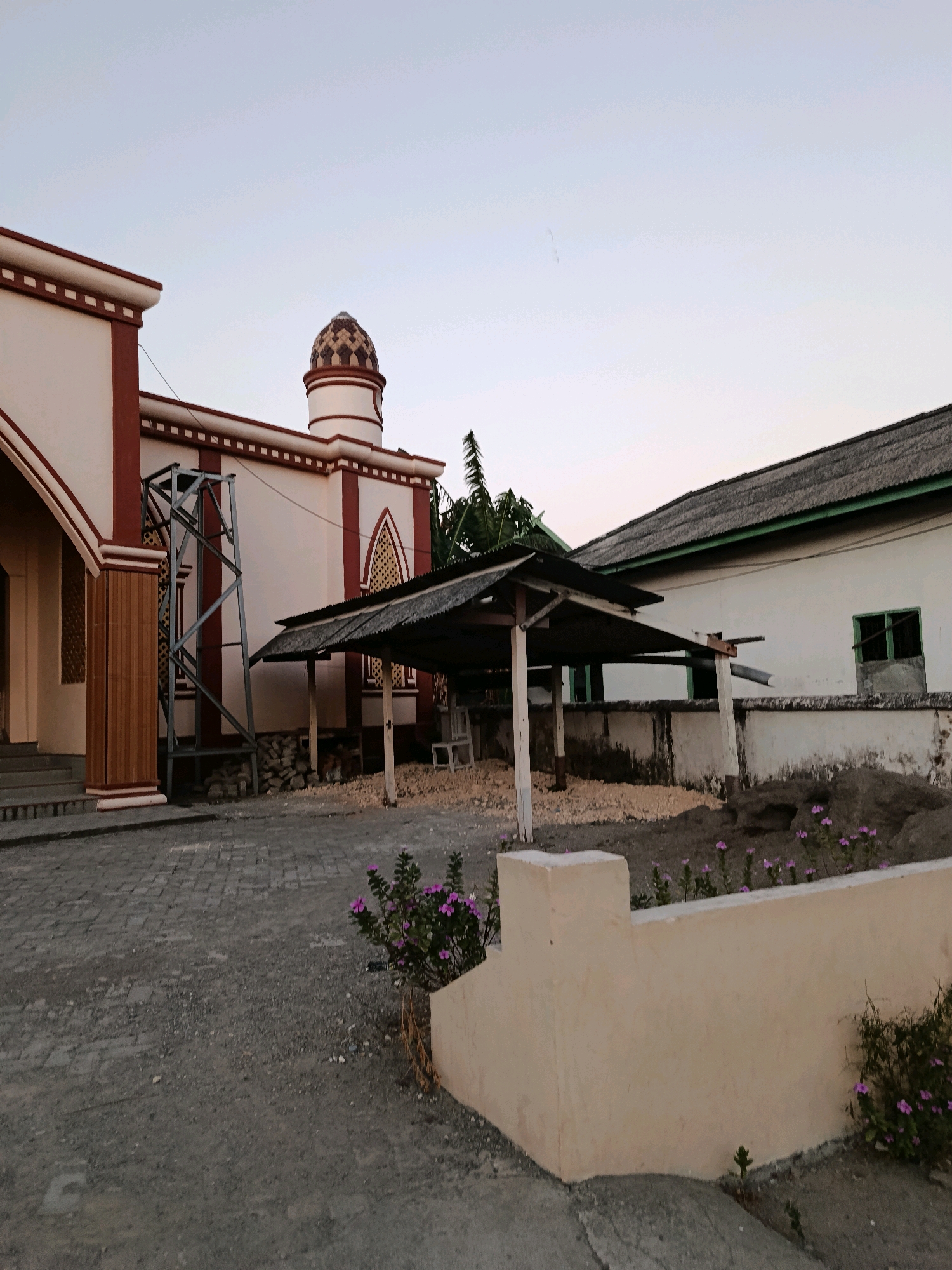 The image shows the exterior of a building that appears to be a mosque or religious structure in Bulukumba, Indonesia. The building features white walls with maroon trim and a decorative dome. To the left, there's an entrance with stairs and wooden pillars. In the center, a metal support structure stands next to a covered outdoor area with a corrugated metal roof. To the right, another building with a dark, textured roof and white walls is visible, featuring a green-framed window. The ground is paved with dark cobblestones, and there are patches of gravel and dirt with some small purple flowers. The sky is clear and pale, suggesting it might be early morning or late afternoon. No people are present in the image. The overall scene depicts a section of a religious complex in a somewhat arid or undeveloped environment.