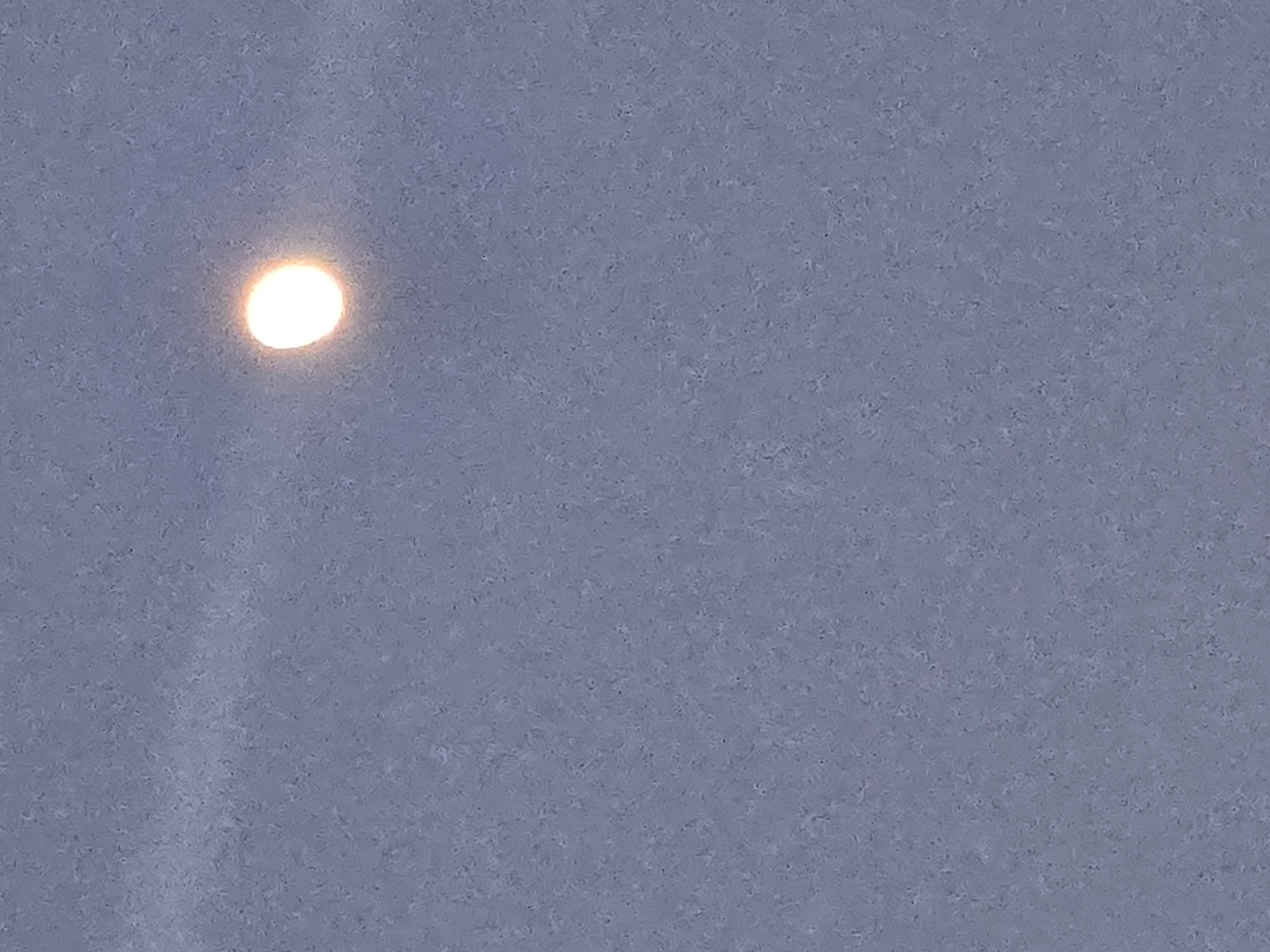 A distant, illuminated orb is positioned at the upper left corner of the frame. The background is a mottled grey-blue tone, which appears uniform throughout the majority of the image. There are no other visible objects or subjects. The location is not discernible from the image.