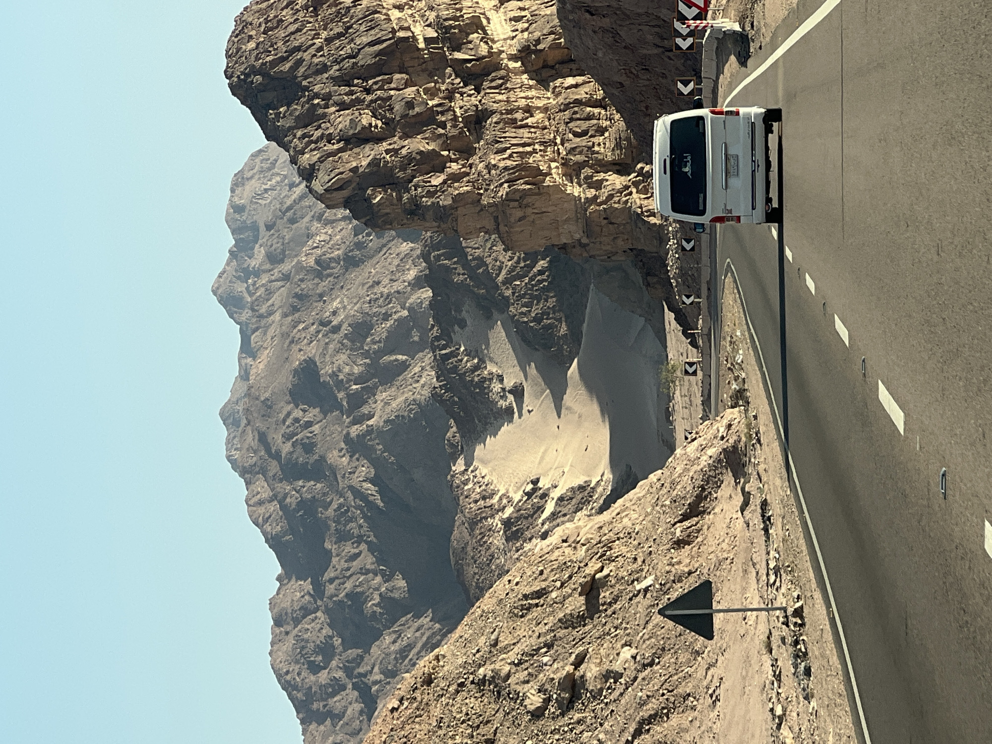 The image shows a white minivan driving on a two-lane asphalt road through a mountainous desert landscape. The road is flanked by rugged, rocky terrain with sparse vegetation. In the distance, large mountains with layered rock formations dominate the horizon under a clear blue sky. The minivan is positioned in the center of the frame, traveling away from the viewer. Road signs indicating curves and potential hazards are visible along the roadside. The lighting suggests it is daytime, likely morning or afternoon, with sunlight casting shadows on the mountains. This scene is consistent with the desert roads found in and around Dahab, Egypt, known for its mountainous terrain and arid environment.