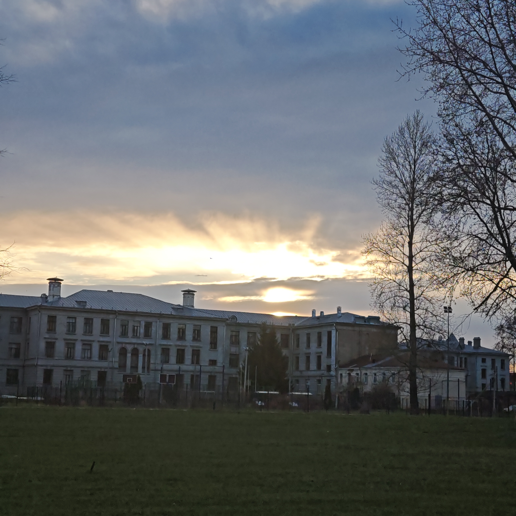 The image depicts a large, grey, multi-story building with a flat roof and multiple windows, situated behind a green lawn. The building appears to have architectural features such as chimneys and a section of the roof is slightly elevated. A fence runs along the edge of the lawn, separating it from the building. Bare trees are visible on the right side of the frame. The sky is overcast with a bright, yellow-orange glow around the horizon line, suggesting either sunrise or sunset. There are no people visible in the image. The location is Unknown, Unknown.