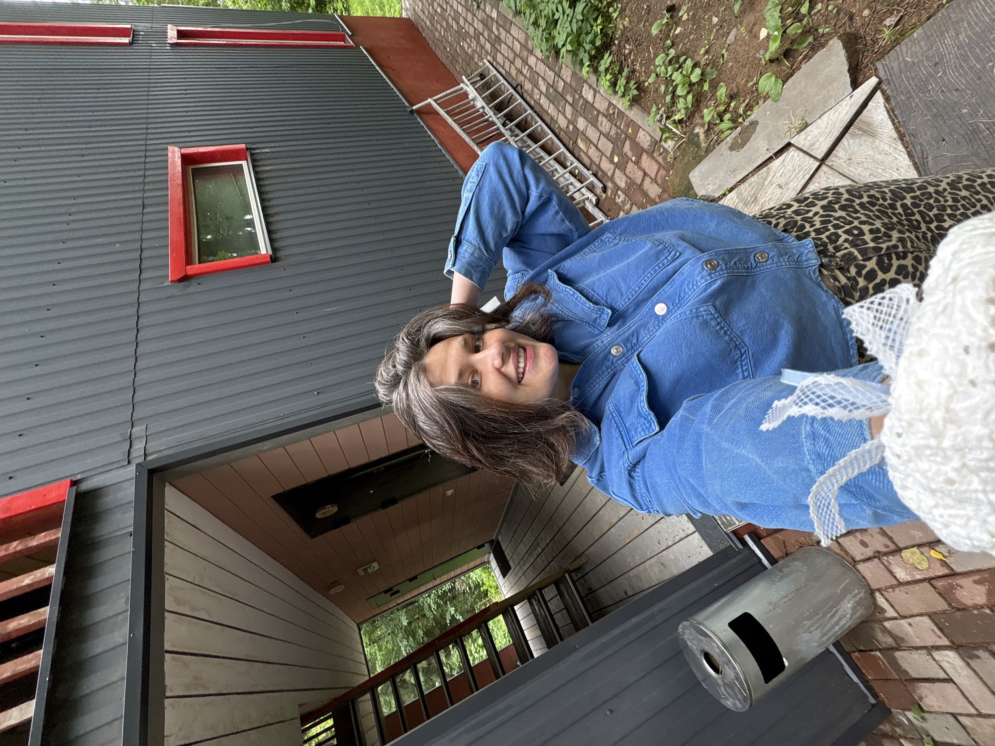 A woman with graying brown hair and a denim jacket smiles at the camera, holding a white object wrapped in netting. She is standing on a brick pathway next to a modern, angular building with dark gray corrugated metal siding and red accents around windows and doors. To her left, an external metal staircase leads up to a balcony. A silver trash can is visible near the staircase. In the background, green trees suggest a natural setting. The scene appears to be outdoors.