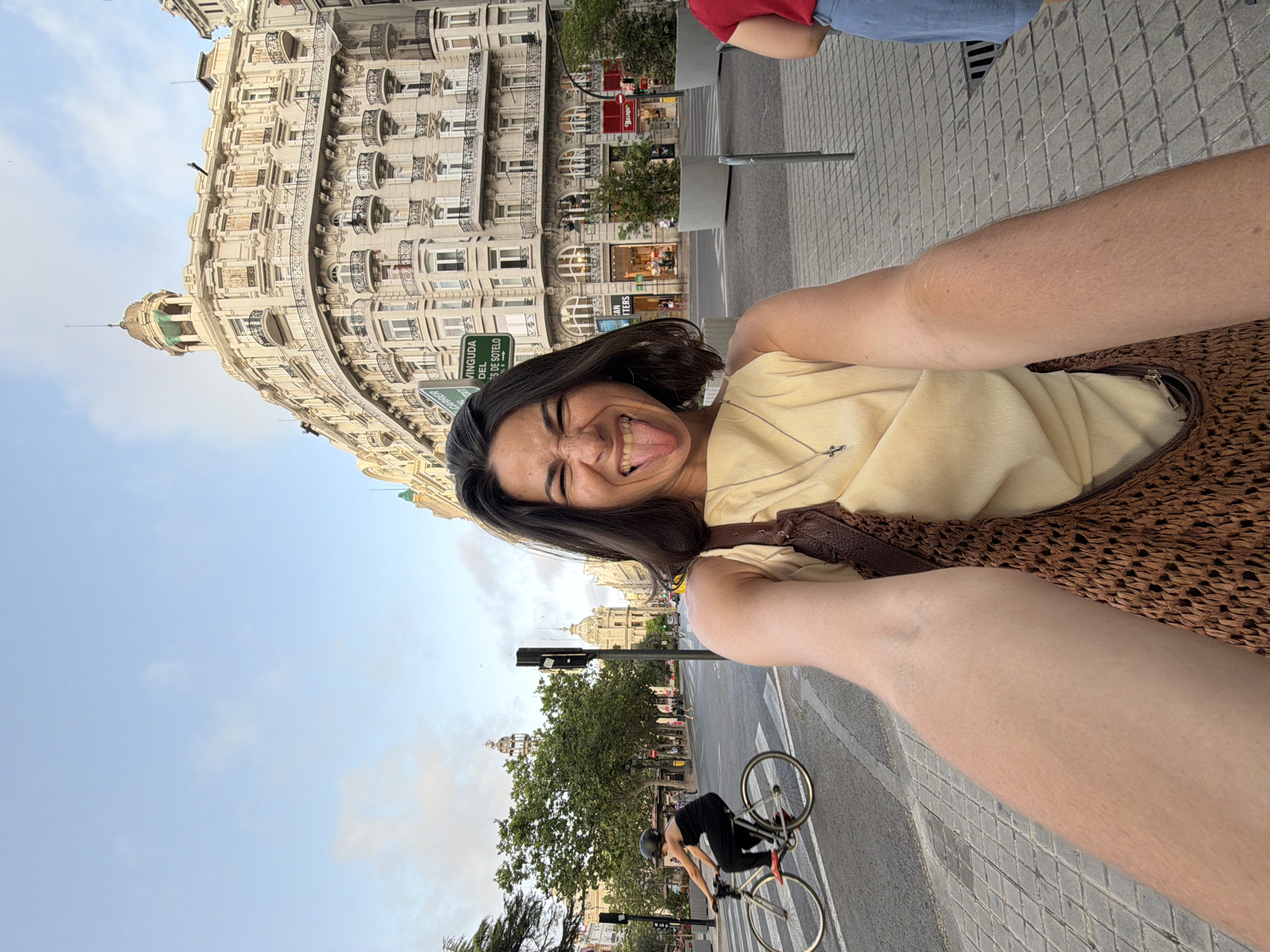 A first-person, close-up selfie of a woman with dark hair and her tongue sticking out, wearing a light yellow top and a brown woven bag. She is on a city street with a cyclist in the background and a large ornate building behind her. The sky is cloudy and blue. The street is paved with gray bricks. There are trees and traffic lights. The building has many balconies and windows.