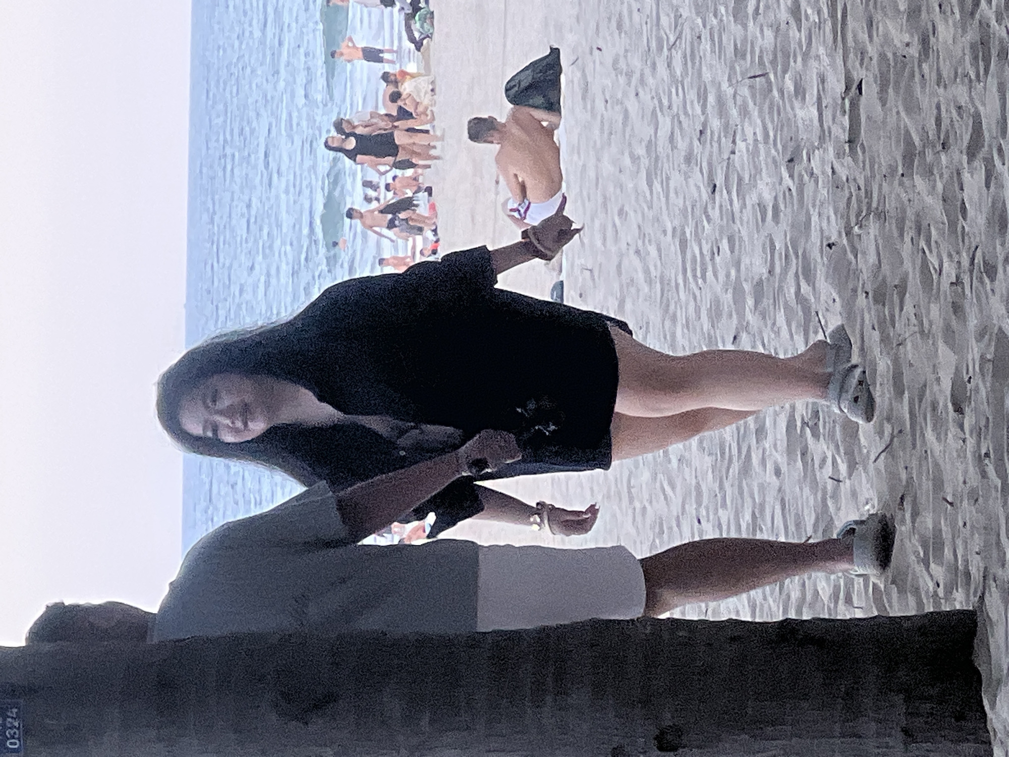 A group of people are on a sandy beach in front of the ocean. The foreground features a man and a woman. The man, partially obscured by a tree trunk on the left, is wearing a grey t-shirt and white shorts. The woman, walking towards the right, is wearing a dark, unbuttoned shirt over a darker garment and short shorts. She is looking to her right and smiling. Her left hand is holding a black object. Both are wearing casual footwear. In the mid-ground, several other individuals are visible, some swimming in the ocean, others walking on the beach. One man is lying on his back on the sand, wearing white swim trunks. The water is blue with some waves. The sky is overcast, suggesting it might be late afternoon or early morning. The scene is likely taking place on My Khe Beach in Đà Nẵng, Vietnam, a popular tourist destination known for its long stretches of sand and clear waters.