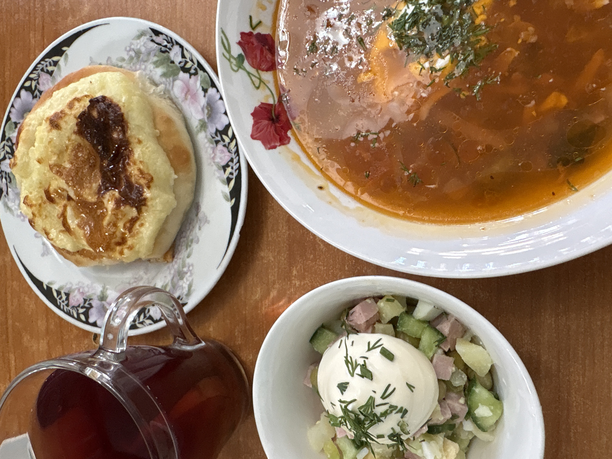 A table setting in what appears to be a restaurant or home, possibly in Bol'shoi Mashlyak, Russia, given the prompt's context. Four main elements are visible: a pastry on a floral-patterned plate, a bowl of soup, a bowl of salad topped with a white sauce, and a glass of dark red liquid. The pastry is round with a slightly browned topping. The soup, garnished with herbs, appears to be broth-based with some solids. The salad is a mix of chopped ingredients topped with a dollop of white sauce, sprinkled with dill. The glass is clear, showing a dark red liquid and has a handle. The table is a light brown wooden surface. The scene suggests a meal setting.