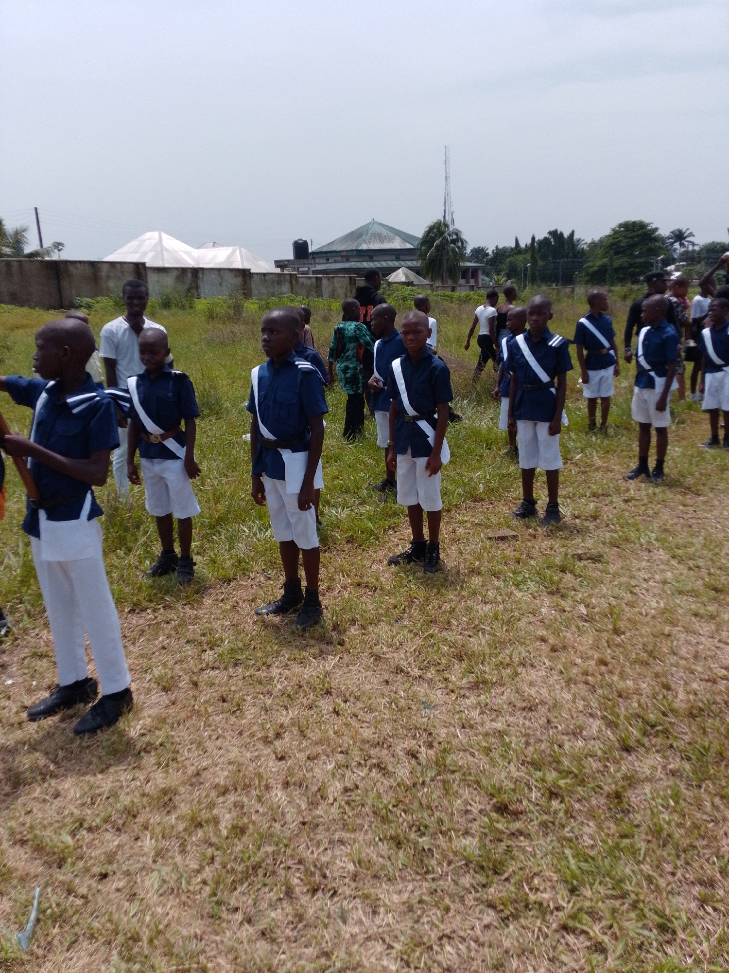 In Abak, Nigeria, a group of boys stand in formation on a grassy field. They are dressed in matching uniforms consisting of dark blue short-sleeved shirts, white shorts or pants, and white sashes worn diagonally across their chests. Black shoes or boots complete their attire. Adults are interspersed among the boys, possibly acting as supervisors or instructors. The setting appears to be an open area, with buildings and trees visible in the background under a partly cloudy sky. A cell tower is visible in the background.