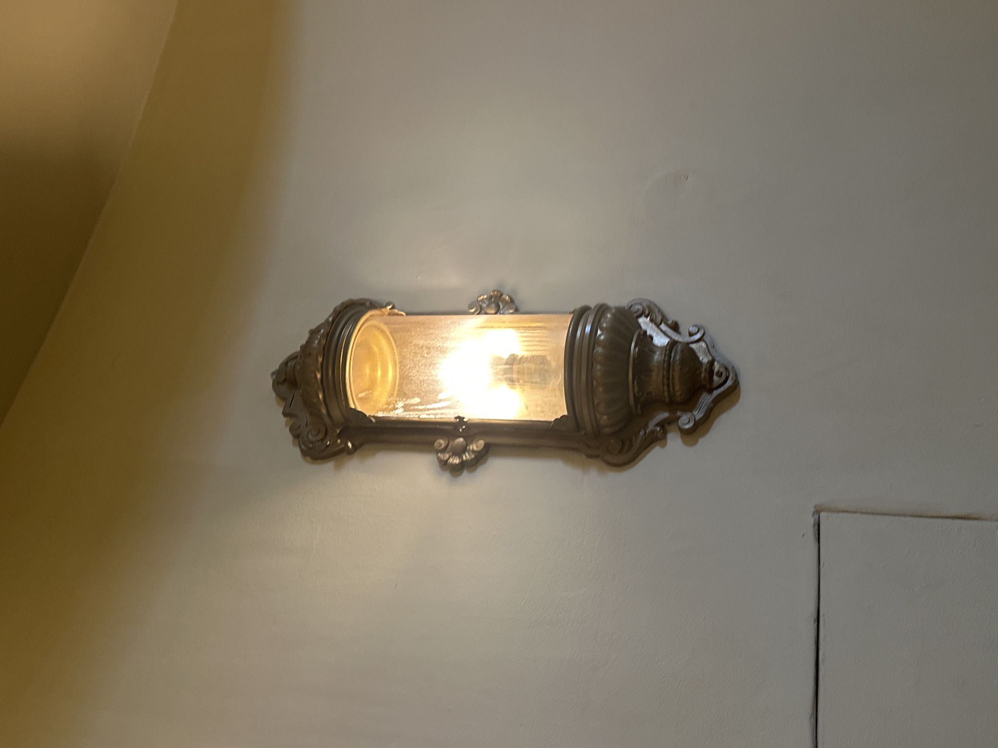 The image shows an antique wall sconce mounted on a textured light-colored wall. The sconce is made of dark, ornate metal and features a clear glass cylinder housing two light bulbs. The light bulbs are illuminated, casting a warm glow through the glass. The wall is painted a neutral tone and meets a darker, possibly painted, ceiling in the upper portion of the image. In the lower left quadrant of the image, a portion of a rectangular architectural element, possibly a vent or a recessed panel, is visible. The lighting suggests an indoor environment. No people or specific Odesa landmarks are visible.