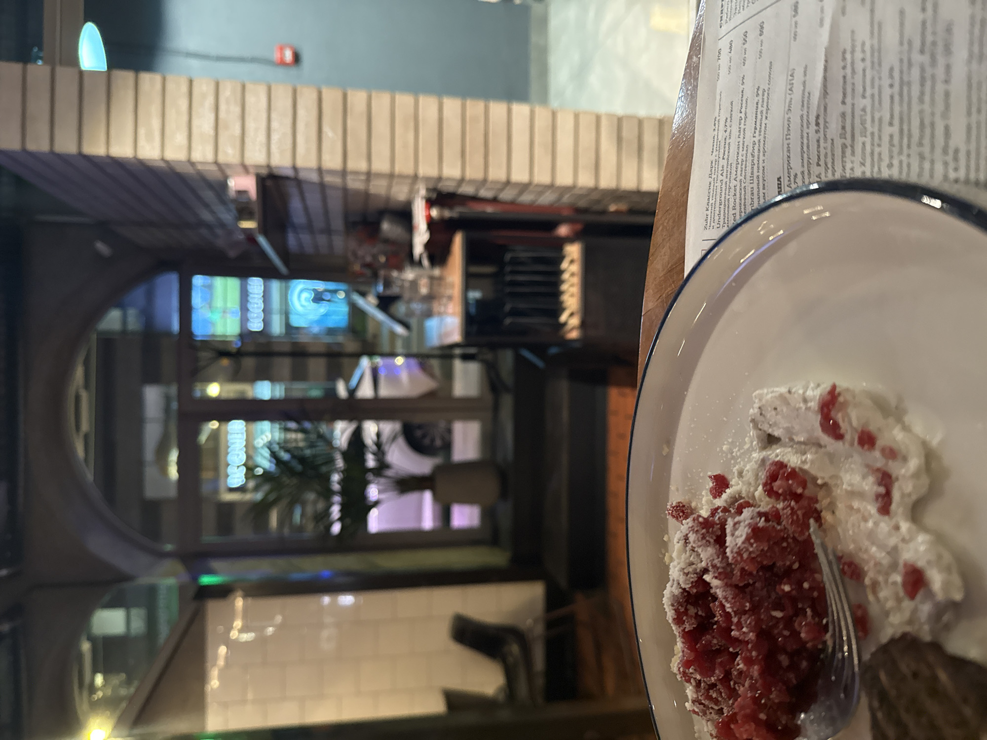 The image depicts a close-up view of a dessert, likely red velvet cake with whipped cream and berries, on a white plate with a blue rim, placed on a wooden table. A fork is partially submerged in the dessert. In the background, a dimly lit interior of a restaurant or cafe is visible. An arched doorway leads to an outdoor or another section of the establishment, revealing a potted plant, a glimpse of a car outside, and neon signs. The architecture suggests an urban setting, possibly St. Petersburg, Russia, given the Cyrillic text on a menu partially visible in the foreground. The overall ambiance is that of an evening meal or outing.